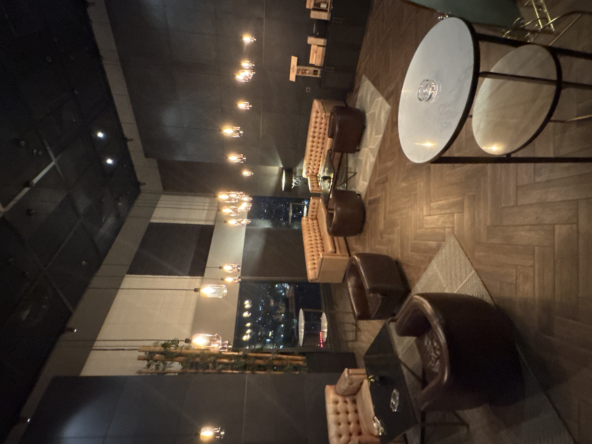 The image depicts an indoor lounge area with a panoramic view of the city at night, likely Sofia, Bulgaria, given the context. The room is furnished with several plush seating arrangements, including a tufted leather sofa and matching armchairs in a warm, light brown hue, and two darker brown, barrel-shaped armchairs. A dark rectangular coffee table sits in front of the light brown sofa, with an ashtray and a small potted plant on it. Another smaller, round marble-topped side table with two tiers is positioned in the foreground, also holding an ashtray.
The walls are dark, with a textured finish. The ceiling is black and features a modern lighting system with numerous pendant lights hanging at varying heights, emitting a warm, ambient glow. Some of these lights are cylindrical glass shades, while others appear to be more decorative clusters of smaller bulbs. A tall, decorative bamboo plant stands in a dark planter near a large window that offers a view of the illuminated cityscape.
On the right side of the room, a dark cabinet with shelves displays several wooden boxes and bottles, suggesting a bar or a display area for beverages. The flooring is made of wooden planks laid in a herringbone pattern. The overall atmosphere is sophisticated and dimly lit, creating an intimate setting. No people are visible in the image.