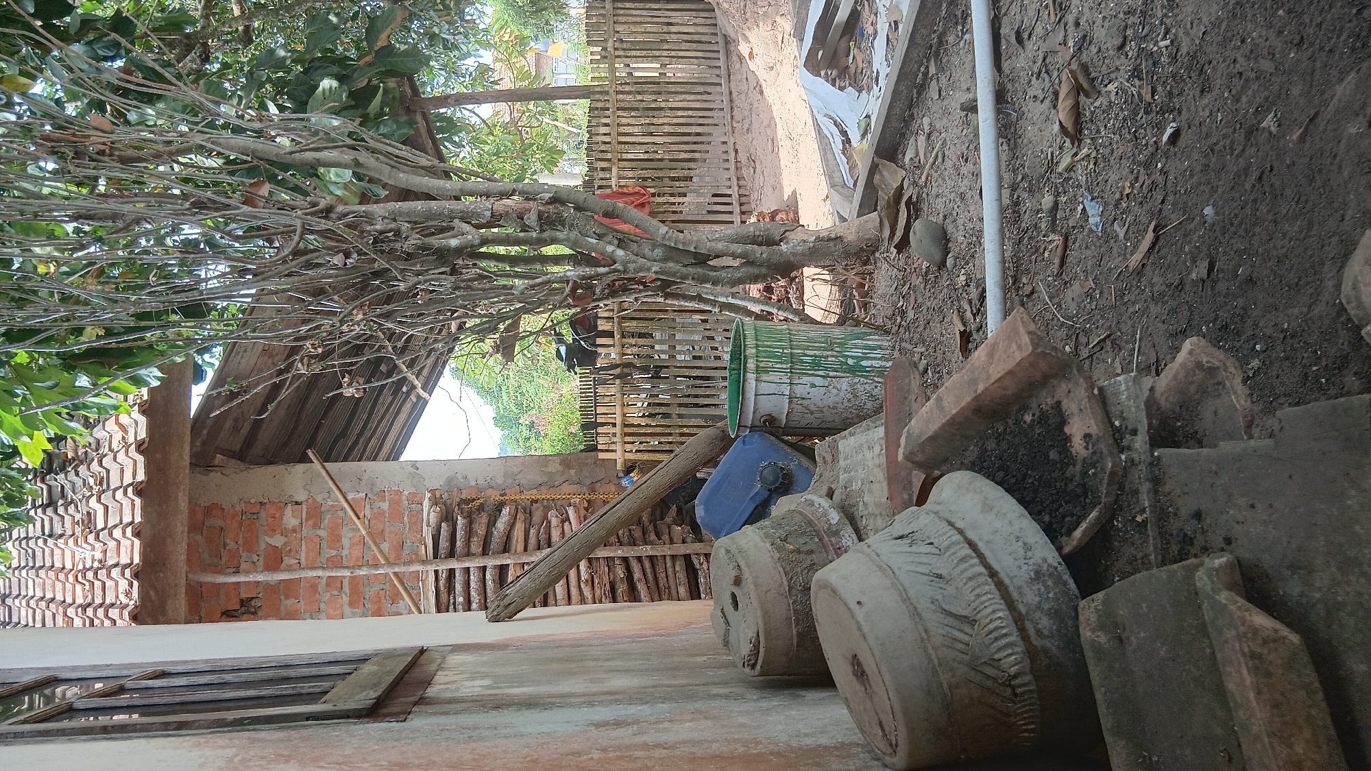 The image depicts a cluttered outdoor area adjacent to a building with a tiled roof and brick walls. In the foreground, several large, weathered ceramic pots are stacked, some containing dark soil. Pieces of broken terracotta tiles and a long, weathered wooden beam lie scattered on the ground. A blue plastic container and a green metal drum with white and green paint streaks are also visible amongst the debris. A thin tree with bare branches stands prominently in the mid-ground. Beyond the immediate clutter, a wooden fence encloses an area with sandy ground. A motorcycle is partially visible behind the fence. In the background, lush greenery and distant hills suggest a rural or semi-rural setting. The overall scene conveys a sense of disarray and disuse. This scene is consistent with a residential backyard or storage area in Prabumulih, Indonesia, a city known for its tropical climate and developing infrastructure, where such elements are commonly found.