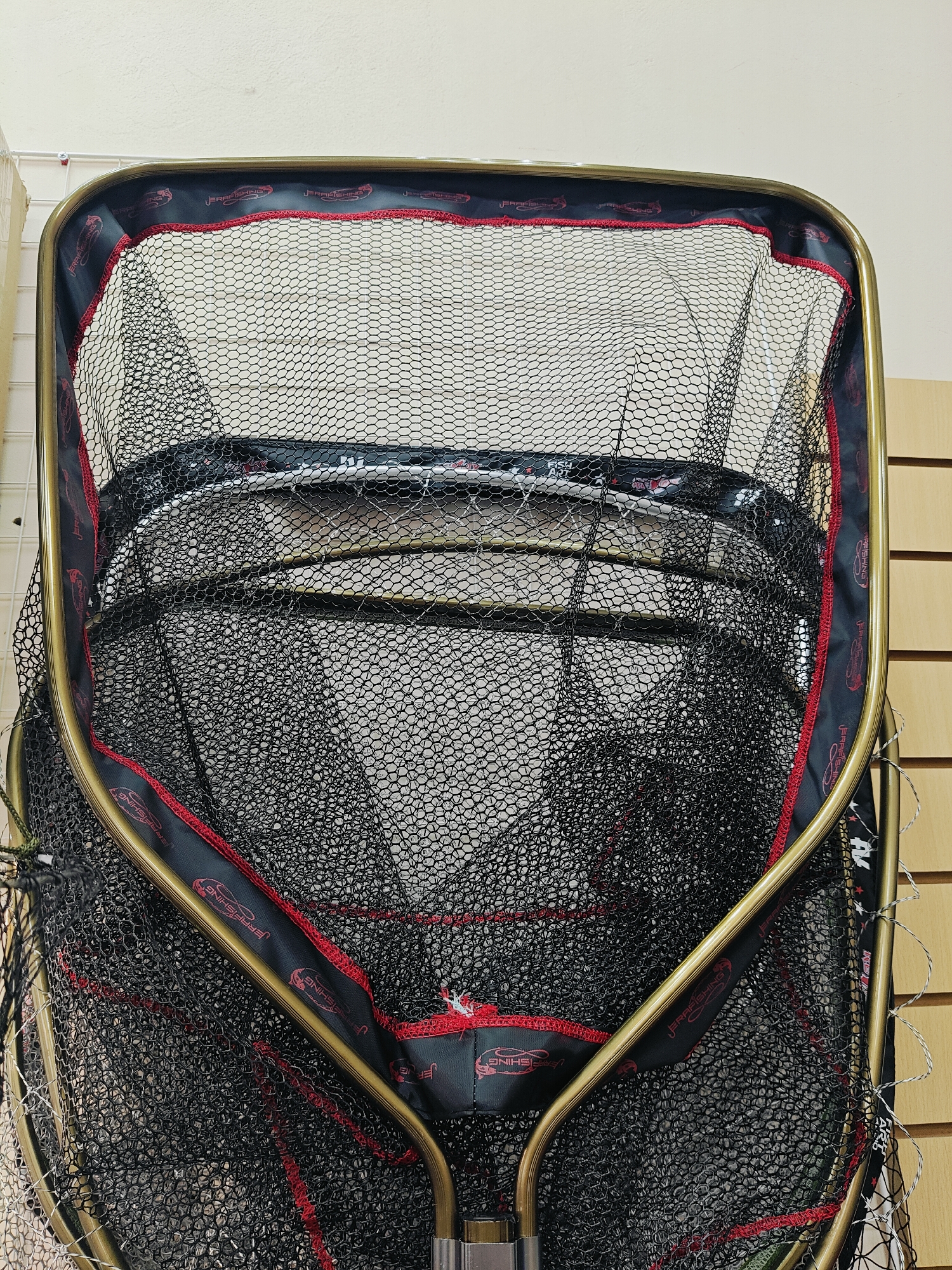 A close-up, high-angle shot shows a black fishing net with a red and black patterned trim, attached to a gold-colored frame. The net is suspended against a white, textured wall with a light brown wooden panel to the right. The pattern on the trim repeats the words "JERRA FISHING" with a fish logo. The net appears to be hanging on a display or in a storage area. The location is the city of Chaikovsky, Russia.