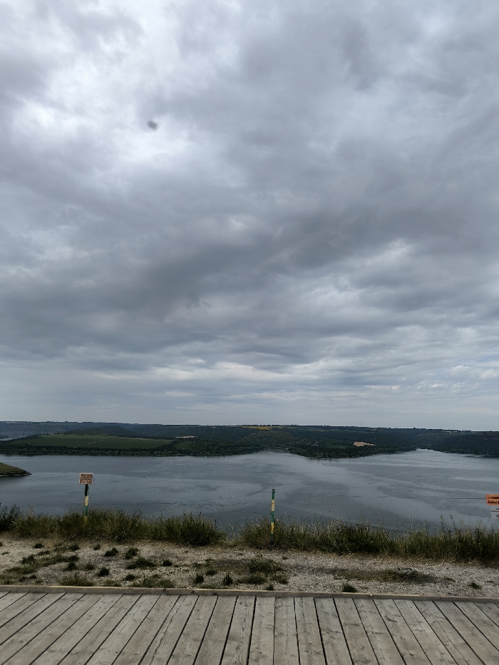 The image presents a panoramic landscape captured from an elevated position in Horaivka, Ukraine. The upper half of the frame is filled with a dense, uniform layer of gray overcast clouds, featuring a distinct dark circular spot in the upper-left quadrant. The mid-ground is occupied by a wide, calm body of dark gray water, likely a river or reservoir, which reflects the somber sky. This water feature is bordered by distant landmasses, displaying undulating terrain covered by a mix of dark green dense vegetation and lighter, possibly agricultural, fields. The immediate foreground consists of a light-colored wooden deck composed of parallel planks. Beyond the deck, a strip of sparse, dry grass and earth precedes the water's edge. On the left side of the foreground, a brown rectangular sign is partially visible, mounted on a pole with alternating green and yellow stripes. A second, identical green and yellow striped pole is present on the right foreground. No individuals are depicted in the scene.