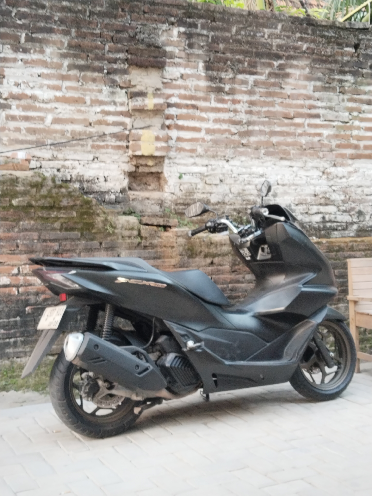 A black scooter is parked on a paved walkway in front of a weathered brick wall. The scooter has a sleek, modern design with sharp lines. A license plate is visible on the rear fender. The brick wall behind the scooter is made of rough, uneven bricks, some of which are covered in moss and grime. The overall scene appears to be an outdoor setting in a tropical or sub-tropical climate, possibly in a courtyard or alley. There are no people visible in the image. The lighting suggests it is daytime.