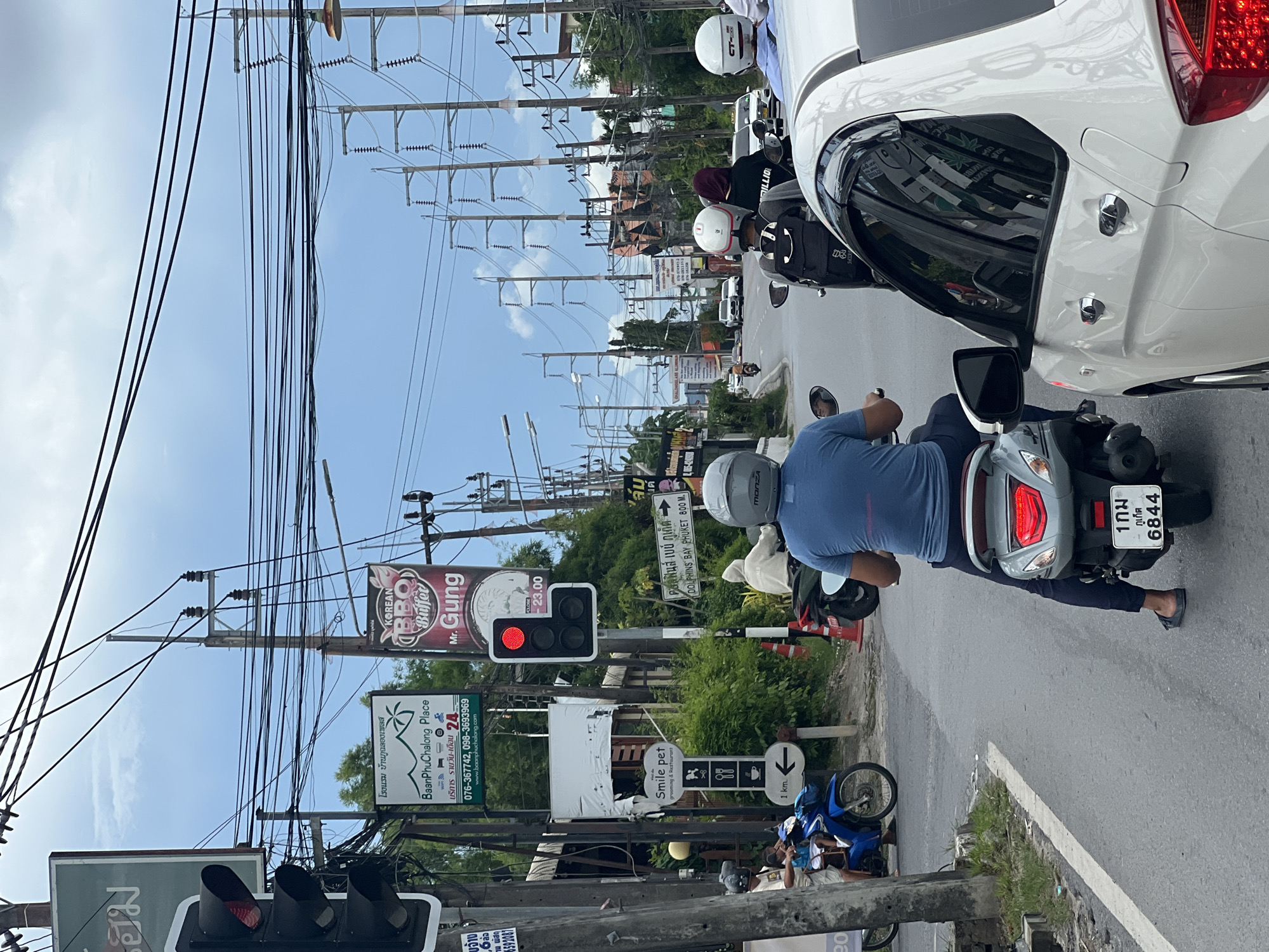 This is a vertical, eye-level shot of a street scene in Chalong, Thailand. The main subjects are people riding motorcycles and a white car. A person in a blue shirt is riding a scooter in the foreground, facing away from the camera. To their right is the back of a white car. In the background, there are several other motorcycles and cars on the road, as well as utility poles with numerous wires, trees, and buildings. The sky is blue with some clouds, suggesting it is daytime and the weather is fair.
Several signs are visible. One sign reads "KOREAN BBQ Buffet Mr. Gung" with a price range. Another sign indicates "BaanPhuChalong Place" and lists services and contact information. A directional sign points to "DOLPHINS BAY PHUKET 800 M." and another points to "1 km." There is also a "Smile pet grooming & Restaurant" sign. A traffic light is visible with a red light illuminated. The license plate on the scooter in the foreground reads "6844". The overall scene depicts a busy street with ongoing traffic and commercial establishments.