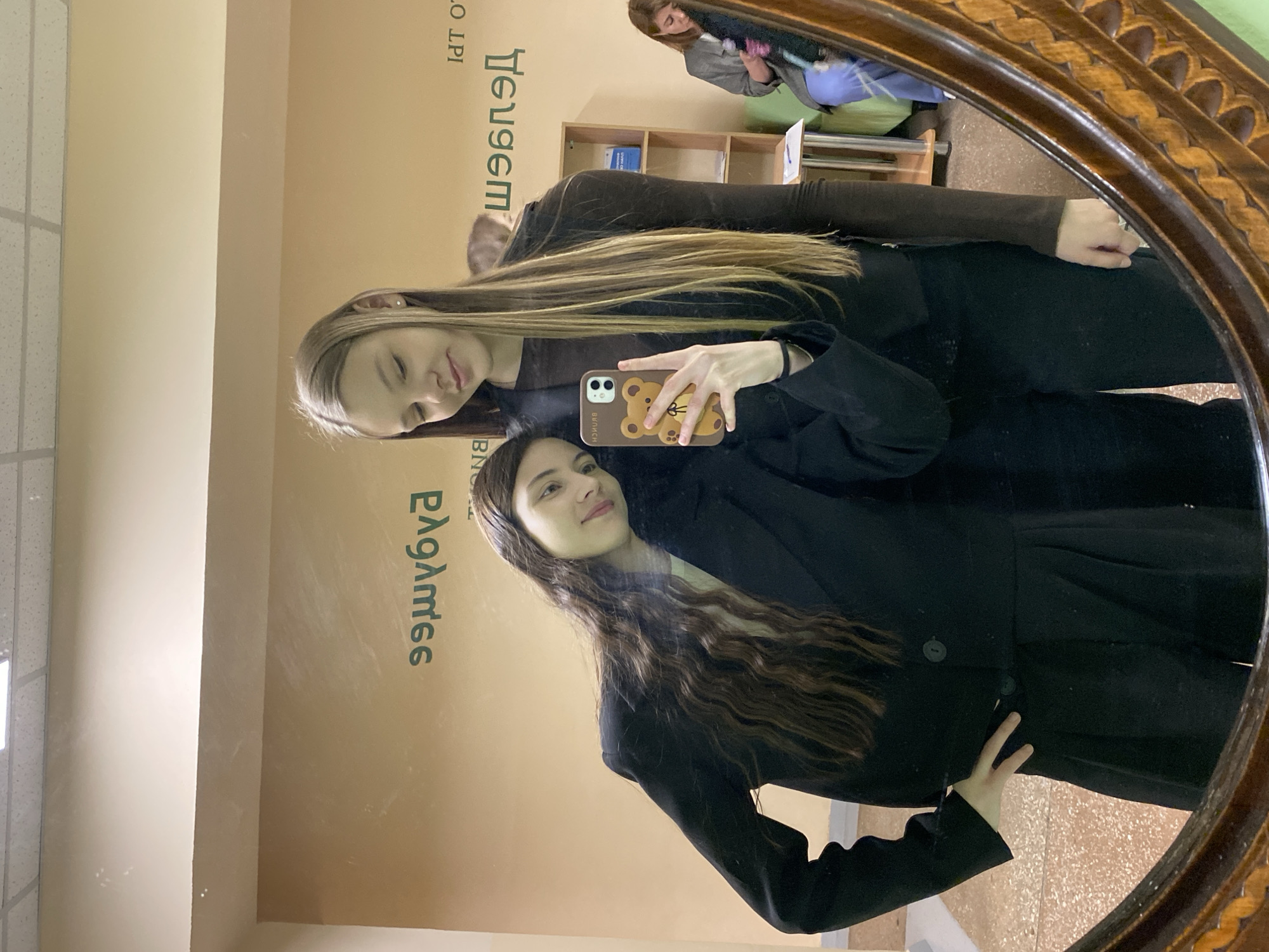 Two young women are taking a selfie in a mirror. The mirror has an ornate wooden frame. Behind them, reflected in the mirror, is a room with light beige walls, and some writing in Russian. There is a bookcase and another person sitting in the background. The location is likely an indoor space in Votkinsk, Russia, given the prompt's instructions. One woman is wearing a black blazer. The other is wearing a brown long-sleeve shirt and holding a phone with a bear-shaped case. They are both facing the mirror.