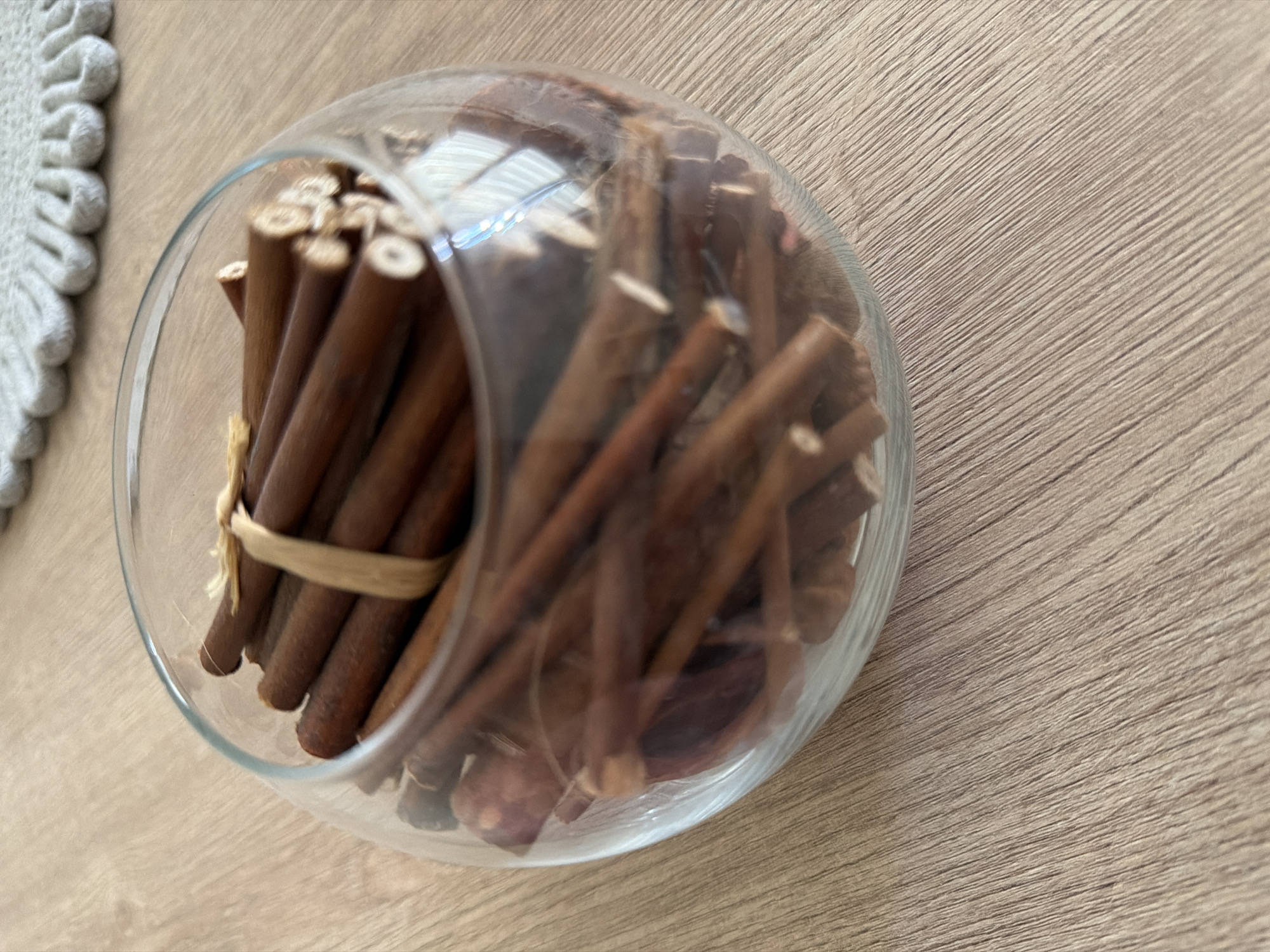 A close-up shot shows a clear glass bowl filled with cinnamon sticks. The bowl is placed on a light brown wooden table. Some of the cinnamon sticks are tied together with a thin, light brown string. The background is slightly blurred, showing a textured grey surface and a sliver of what appears to be a rug with a scalloped edge. The lighting suggests an indoor setting, possibly a home or a decorative display. The scene does not depict any specific urban location within Pazardzhik, Bulgaria, or any actions or interactions. It is a still life composition focused on the cinnamon sticks in the bowl.