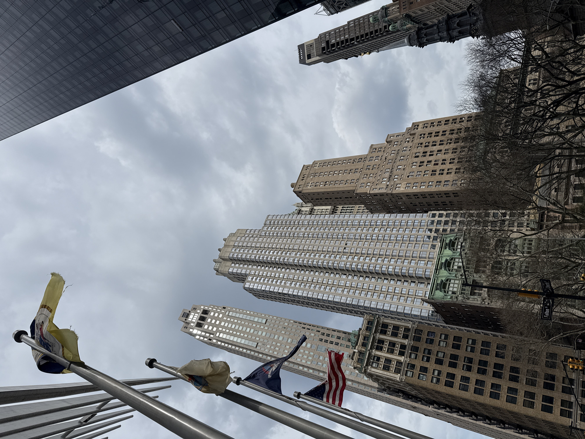 The image presents a low-angle view of a cluster of buildings against a cloudy sky in New York City, USA. In the foreground, several flags on poles are visible, including an American flag and what appears to be a New York State flag. The surrounding architecture is typical of the city's dense urban environment, with a mix of older and more modern skyscrapers. The perspective emphasizes the height and scale of the buildings. The image seems to capture a moment in the Financial District of New York City.
