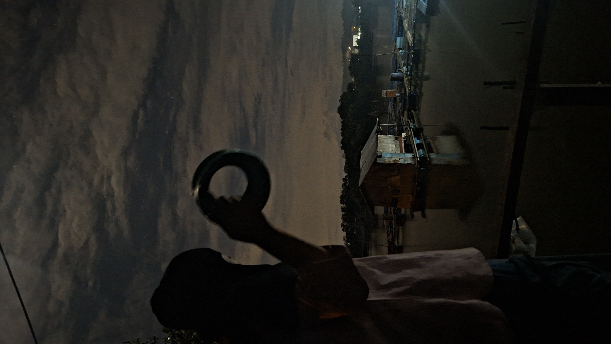 A silhouette of a person, wearing a dark head covering and a light-colored shirt, stands on the left side of the frame, holding a circular object, possibly a Tambourine or a ring-shaped toy, up to their face. The person is looking towards a body of water at night. In the water, there is a small, dilapidated wooden structure, likely a floating house or fish farm shack, with a corrugated metal roof. Around the shack, various floating structures and possibly boats or rafts are visible, indicating a village or settlement on the water. The background shows a dark silhouette of trees and a distant, dimly lit cityscape with lights suggesting the city of Lhokseumawe, Indonesia. The sky is dark with scattered clouds, and a few faint stars are visible. The overall mood is nocturnal and somewhat mysterious.