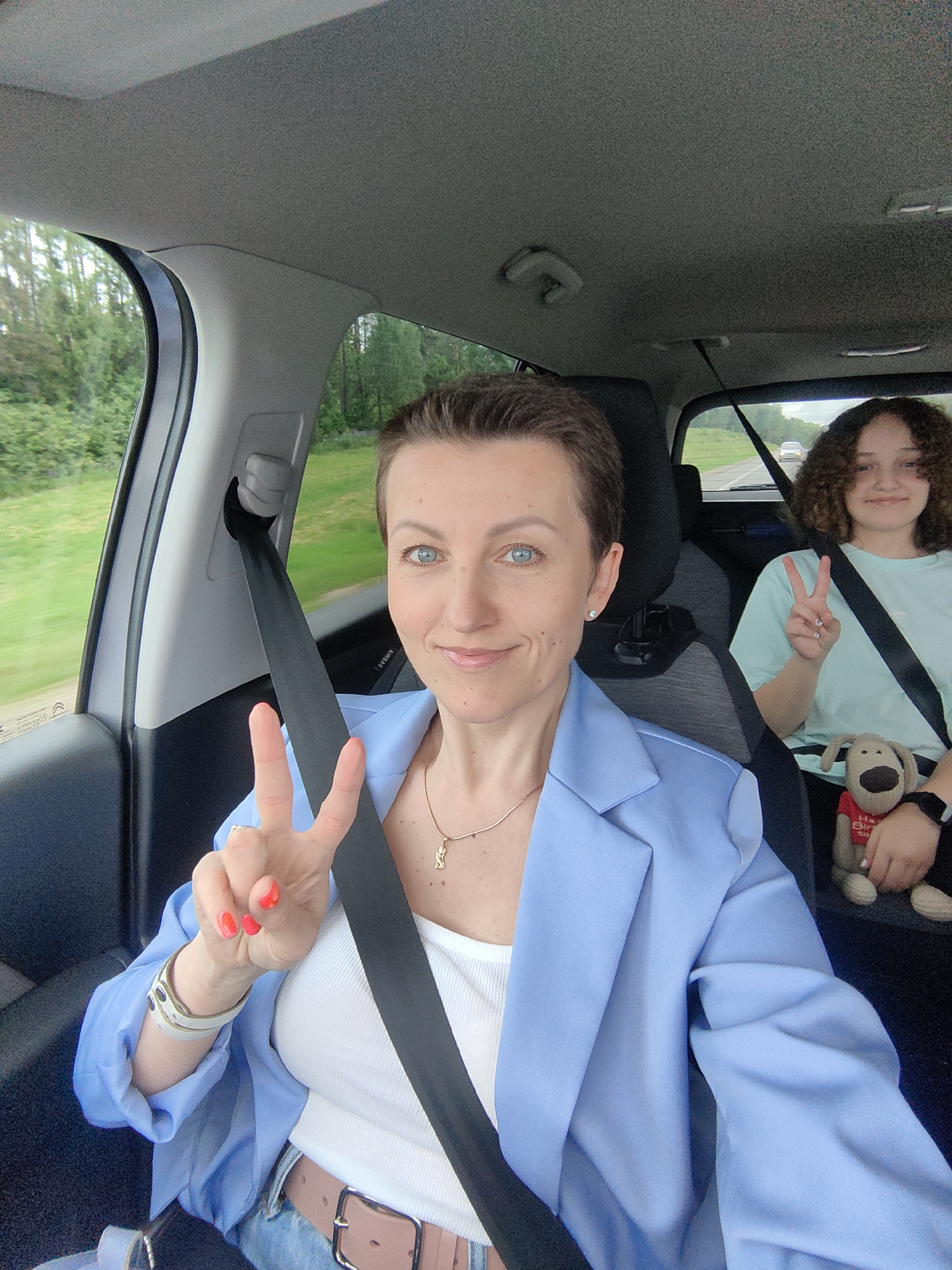 The image shows the interior of a light-colored vehicle in motion, capturing two females. The scene is located in the Дзяржынскі сельскі Савет area of Belarus, evident by the green trees and natural landscape visible through the vehicle's windows, indicating movement on a paved road where another white vehicle is seen in the distance.
In the foreground, a woman with short dark hair and blue eyes, appearing to be in the front passenger seat, smiles directly at the camera. She is wearing a white ribbed tank top under a light blue blazer and is secured by a black seatbelt. Her left hand, adorned with red nail polish and a bracelet, is raised in a peace sign (V-sign). A gold necklace with a small, stylized pendant hangs around her neck.
In the background, a younger female with curly brown hair is seated in the rear, also smiling and making a peace sign with her right hand. She is wearing a light blue or mint green t-shirt and is also secured by a black seatbelt. Next to her, a brown plush dog toy wearing a red t-shirt with "Happy Birthday" text is visible, also belted in.
The vehicle's interior features light grey ceiling upholstery, dark seat headrests, and a visible "AIRBAG" label near the front passenger window. Motion blur in the background further confirms the vehicle is actively traveling.