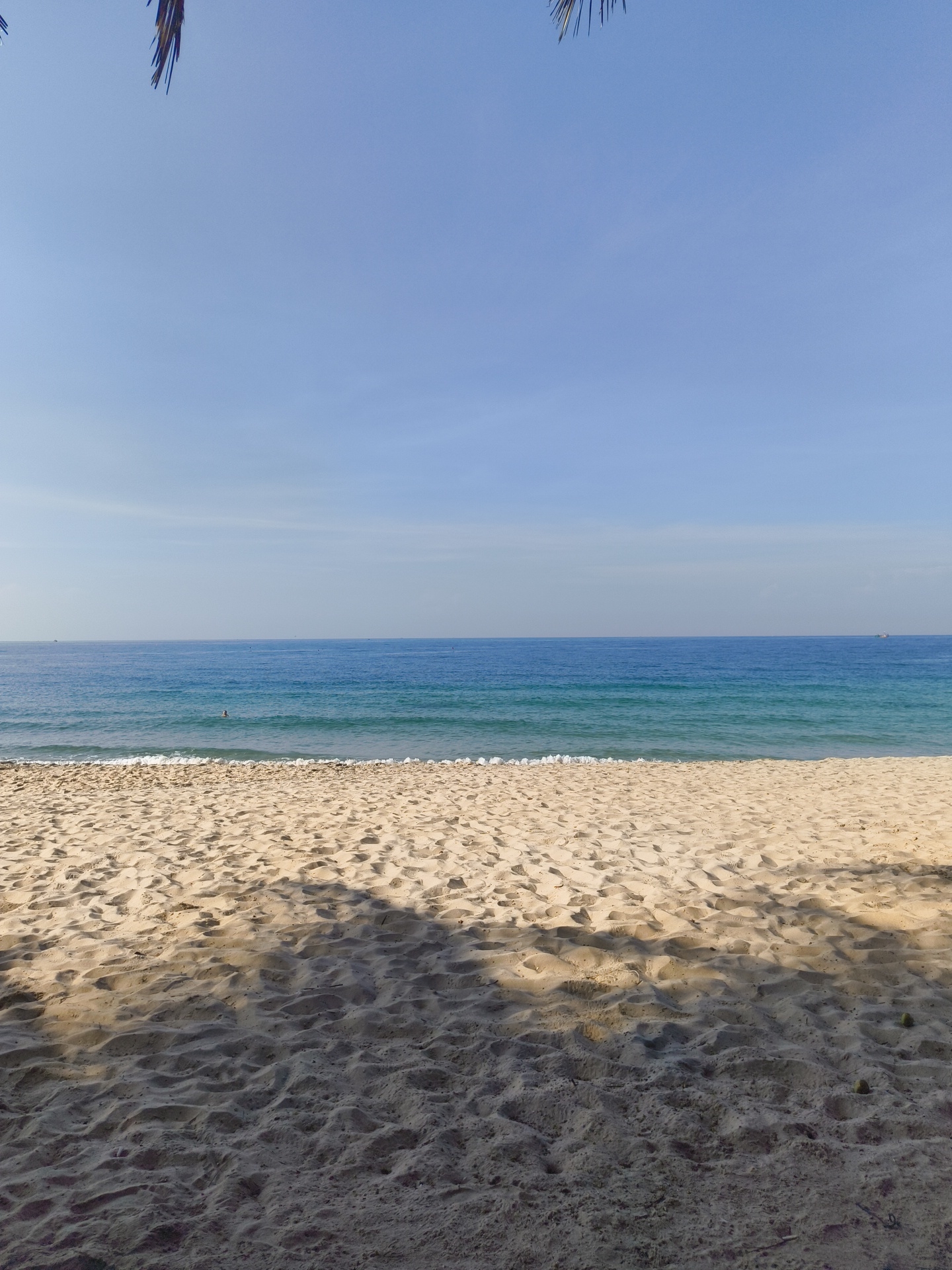 The image depicts a serene beach scene in Phu Thuy Ward, Vietnam. The foreground is dominated by textured, light-colored sand, showing distinct patterns of footprints and subtle shadows cast from an unseen overhead object, likely palm fronds as indicated by the top edge of the frame. The middle ground features a gentle shoreline where small, white waves are breaking. Beyond the breaking waves, the sea transitions from a lighter turquoise near the shore to a deeper, rich blue as it extends towards the horizon. A single person is visible in the water, appearing to swim or float. The horizon line is clear, separating the sea from a pale blue sky with faint, wispy clouds. The overall impression is one of tranquility and natural beauty on a bright, clear day.
