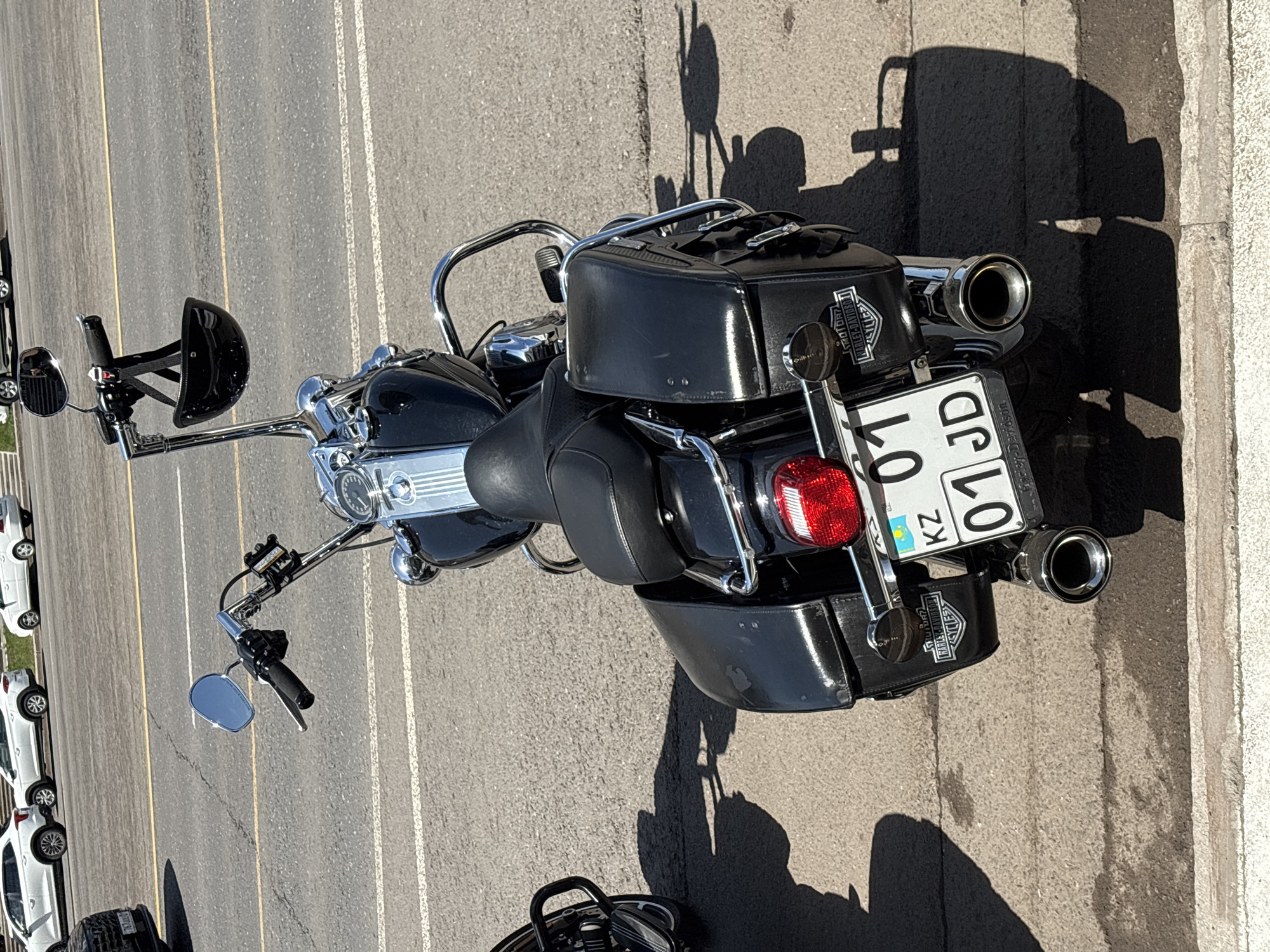 This is a close-up, eye-level, outdoor, daytime, medium shot of a black Harley-Davidson motorcycle parked on a concrete sidewalk in Astana, Kazakhstan. The motorcycle is angled slightly away from the camera, showing its rear and left side. It features black leather saddlebags, chrome accents, a black seat, and a license plate with "KZ" and "01 JD" visible. A small, black helmet is hanging from the handlebars. In the background, a road with white lane markings and several white cars are visible. The light suggests it is a sunny day, casting shadows on the ground.