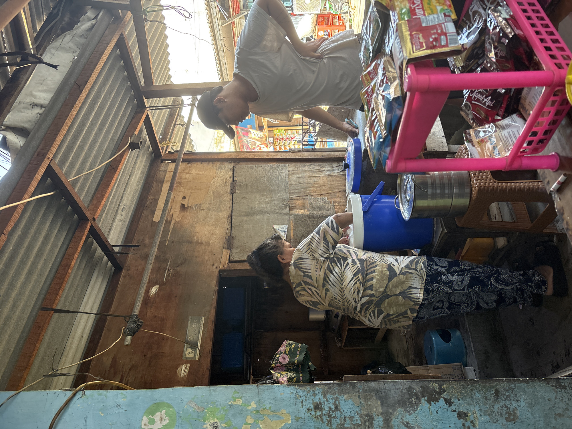A full shot shows an outdoor stall, likely in North Jakarta, Indonesia, under a corrugated metal roof supported by wooden beams. Two individuals are visible. On the left, a person wearing a patterned short-sleeved shirt and dark patterned pants stands facing a large blue insulated container, with their hands inside it. To the right, a person wearing a white t-shirt and a dark baseball cap stands with their left hand on their hip, looking towards the left side of the frame. In the foreground, a pink plastic shelf unit is laden with packaged goods, appearing to be food items or condiments. The stall's structure is made of weathered wood, and various objects, including a blue stool and metal containers, are scattered around. The overall impression is of a simple, functional marketplace or roadside stall.