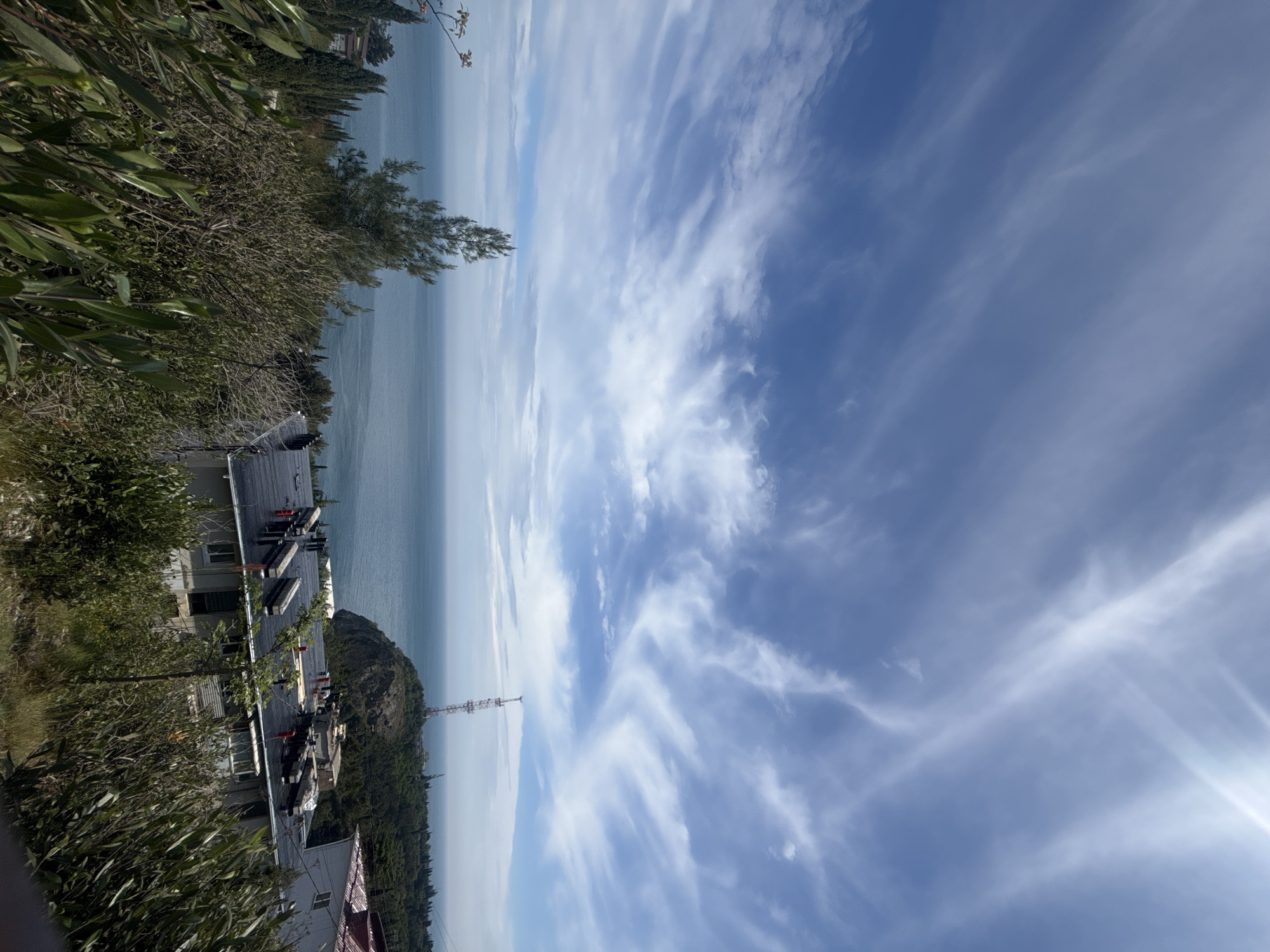 The image depicts a scene with a body of water in the background, possibly a sea or large lake. Above the water, there is a clear blue sky with wispy white clouds. In the foreground, there is a building with a dark roof and several structures on top, possibly chimneys or ventilation systems. Surrounding the building is a mix of green vegetation, including trees and shrubs. A tower-like structure is visible in the distance, potentially a communication tower. The location appears to be a coastal area, with buildings built on a hillside overlooking the water. No people are visible in the image. The photograph is taken from a slightly elevated perspective, and the composition emphasizes the relationship between the natural environment (water, sky, and vegetation) and the built environment (buildings and tower).