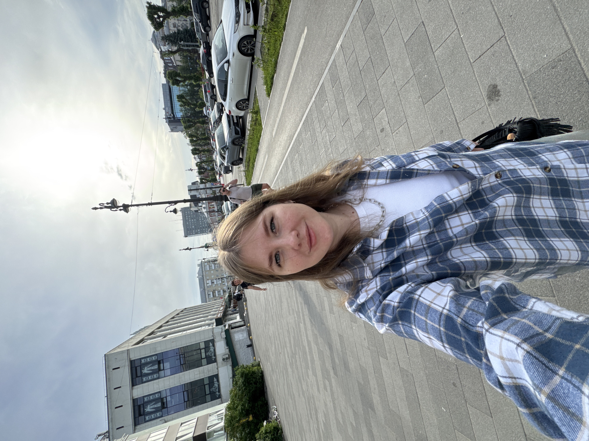 A young woman is taking a selfie on a paved walkway in Perm, Russia. She has fair skin, blond hair, and wears a white tank top under an open blue and white plaid shirt. Buildings line the street behind her, including a light-colored building with repeating motifs in its windows and a taller office building. Cars are parked along the street beside the walkway. A lamppost is visible near a crosswalk. Other pedestrians can be seen in the background. The sky is overcast.