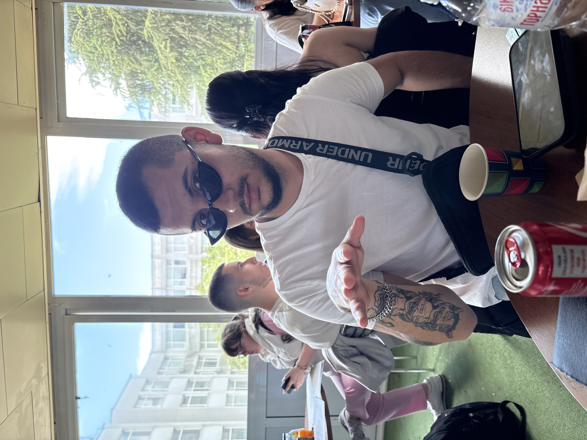 The photo shows a man in sunglasses and a white t-shirt with an Under Armour strap, looking directly at the camera with his left arm extended. His left arm has a tattoo. He is seated at a table with a Coca-Cola can and a colorful cup. Behind him are other people seated, including a woman with long black hair and another person in a pink outfit. Through the windows, buildings are visible, suggesting an urban setting. The image appears to be taken indoors near a window with a view of other buildings. The location appears to be Blagoevgrad, Bulgaria.