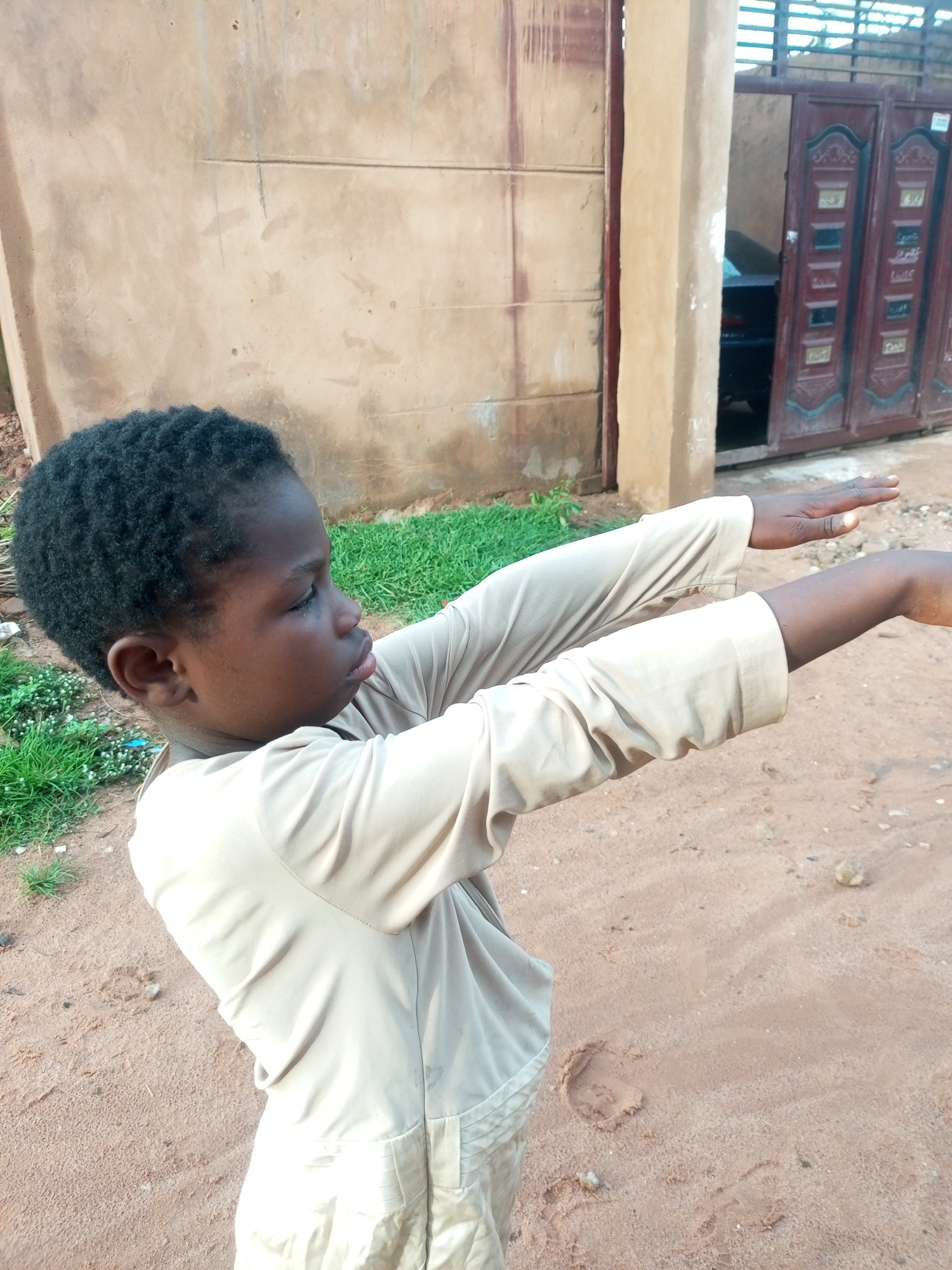 In Tempure, Nigeria, a young Black boy with short, dark, curly hair stands outdoors, facing right and looking slightly upwards. He is wearing a tan, long-sleeved shirt and matching pants. His arms are extended forward and slightly upwards, with his palms facing forward. The background features a textured, light brown building wall to the left, and a dark red metal gate with decorative panels to the right. The ground is sandy and uneven, with patches of green grass visible on the left. The lighting suggests it is daytime, possibly late afternoon.