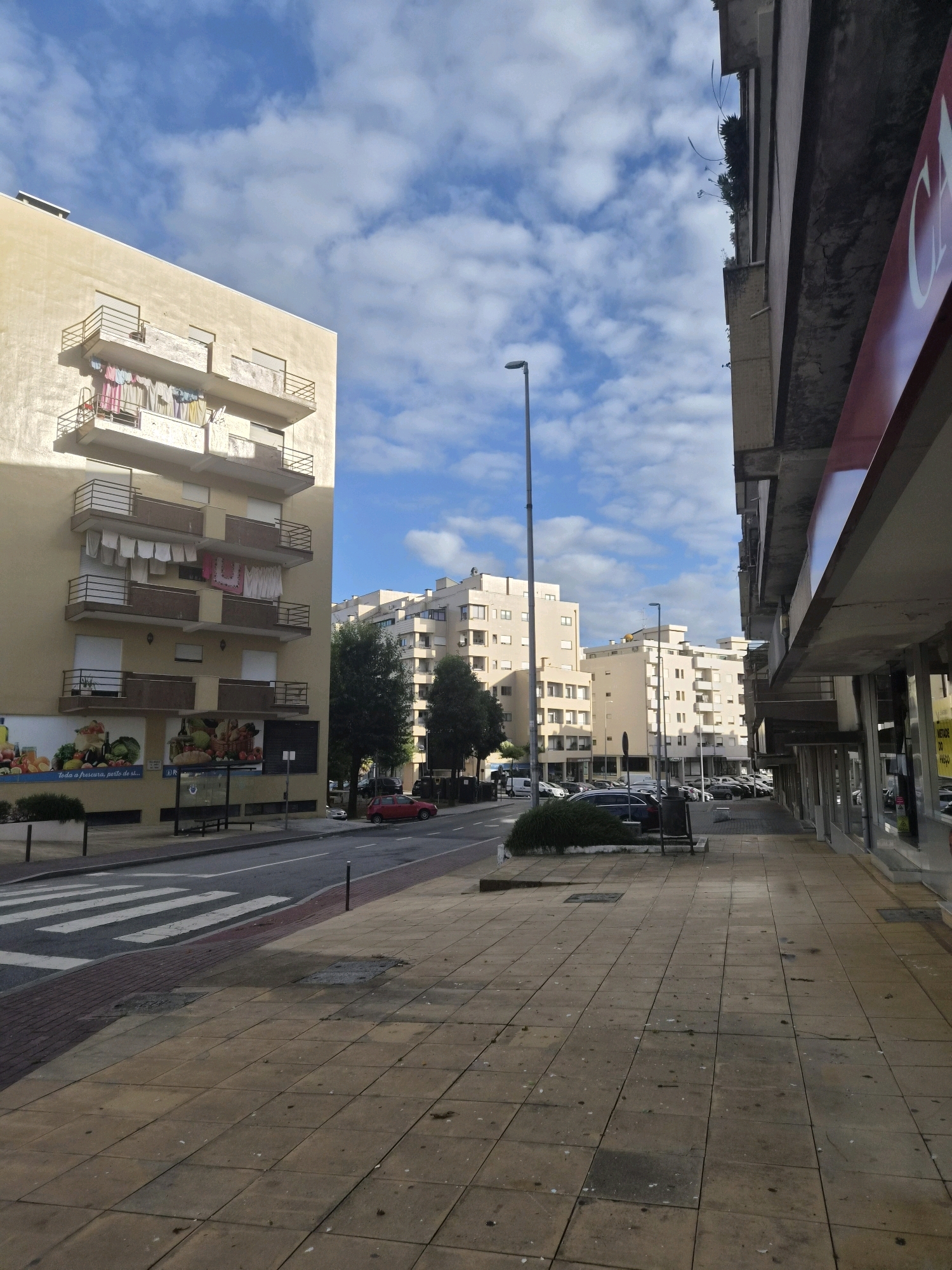 The image depicts a street scene in Barcelos, Portugal, under a partly cloudy sky. On the left, a light yellow multi-story building features several balconies with clotheslines strung across them, displaying laundry. The ground level of this building appears to house commercial establishments, with large advertisements visible. A crosswalk is marked on the road in front of the building. Across the street, more multi-story buildings of modern architectural design are visible. On the right side of the image, the facade of another building with a red awning is prominent, suggesting a commercial space. The sidewalk is paved with square tiles. A street lamp stands on the left side of the road, and another is visible further down the street. A red car is parked on the right side of the road, and several other vehicles are parked along the street on the left. The overall impression is that of a residential and commercial urban area.