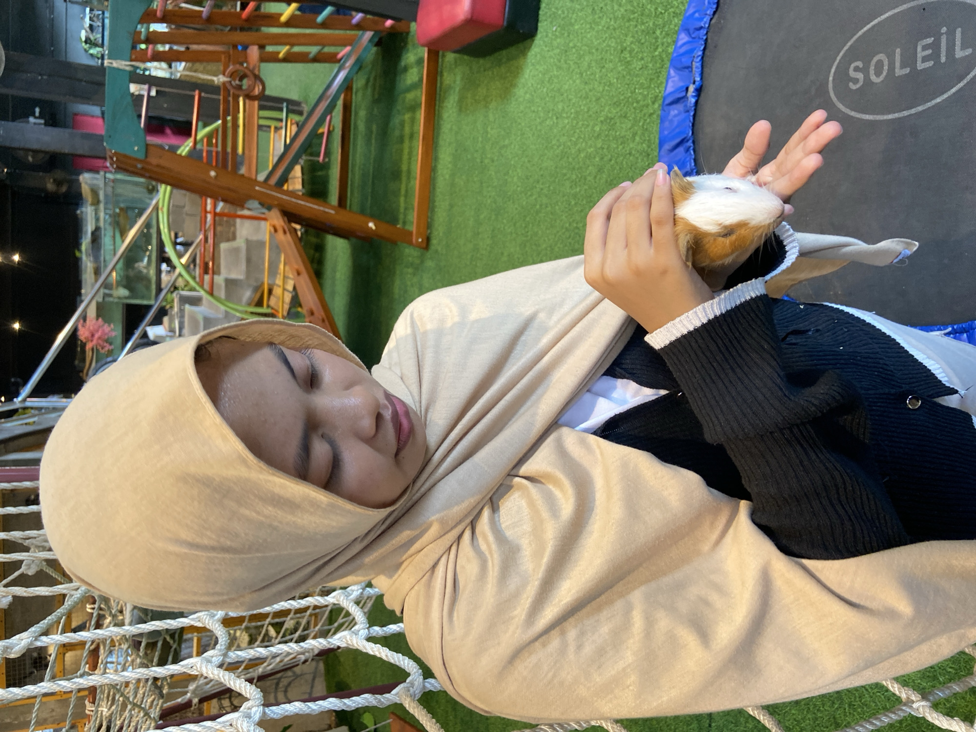 In Rengat, Indonesia, a young woman wearing a beige hijab and a black cardigan over a white shirt is shown holding a guinea pig. The guinea pig is white and brown and is being held in her hands. She appears to be looking down at the animal with a gentle expression. The background features a playground structure with colorful elements and a green turf floor. A trampoline with the word "SOLEIL" printed on it is partially visible in the foreground. A rope net structure is to the left of the woman. The overall setting suggests an indoor recreational area or a pet-friendly establishment.