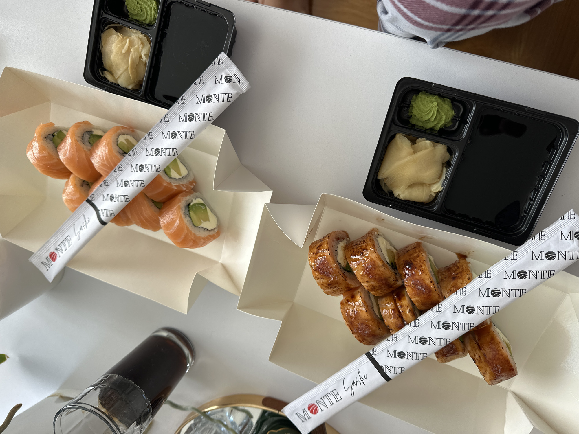 The image depicts a meal of sushi, presumably a takeout order, set on a light-colored surface. There are two white takeaway boxes containing different types of sushi. The box on the left has salmon-covered rolls, while the box on the right contains rolls with a darker, glazed topping. Each box has a wrapped pair of chopsticks with "MONTE Sushi" printed on the wrapper. Accompanying the sushi are two black plastic containers, each containing wasabi and pickled ginger. A glass of dark liquid, likely soy sauce, is also visible, partially obscured in the lower-left corner. The overall setting suggests a casual dining experience, possibly indoors. The chopsticks wrapper includes "MONTE Sushi" that suggests this image was taken near or in a city near Monte such as Budva, Montenegro.