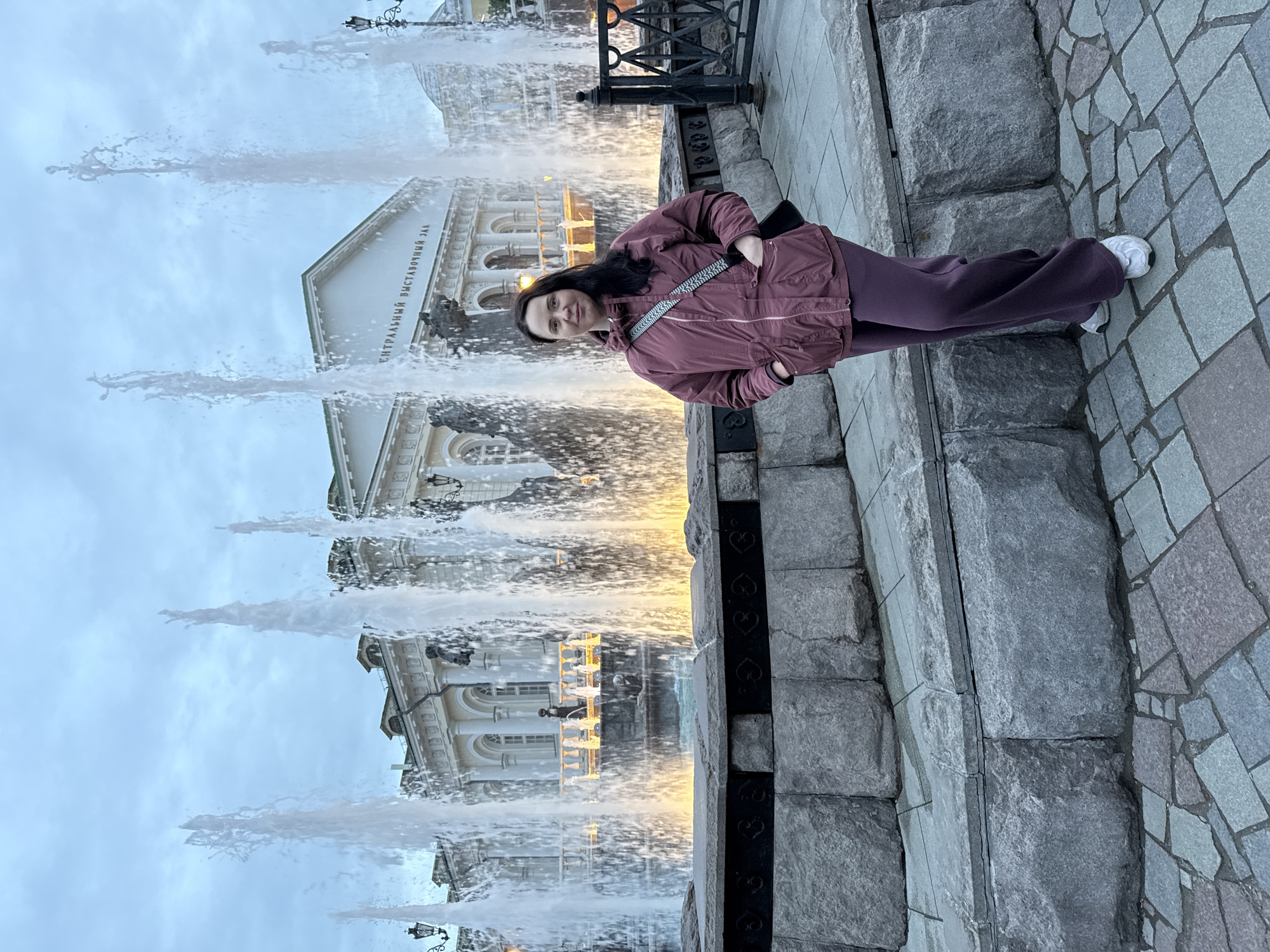 A woman is standing next to the fountain in front of the Manege Square building in Moscow, Russia. The woman is wearing a burgundy jacket and pants with white sneakers. She is smiling and looking at the camera. The fountain has multiple jets of water shooting upwards, and the Manege Square building is visible in the background. The sky is overcast. The woman is standing on stone blocks near an iron fence.
