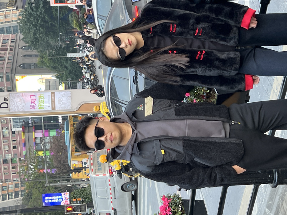 The image captures two East Asian individuals posing in an urban street environment in Yuzhong District, Chongqing, China, specifically identified by a sign indicating "Chaotianmen Delicious Street." The male, positioned on the left, wears dark sunglasses, a grey hooded sweatshirt, and a black and dark grey two-tone jacket with a fleece lower half and nylon upper half, featuring yellow accents and a tan patch on the right chest. He stands with his left hand in his black trouser pocket, looking directly forward. To his right, a female with long dark hair wears narrow black cat-eye sunglasses, a black faux fur jacket with red frog closures and red-lined cuffs, and dark trousers. She has a slight head tilt and gazes towards the viewer's left.
Behind the subjects, a street with moderate vehicle and pedestrian traffic is visible under diffuse daylight. A dark sedan is partially obscured behind the female, while a white van with "AD3910" markings and several delivery scooters, operated by individuals in bright yellow jackets and helmets, are present on the roadway. Pedestrians populate the sidewalks in the background. A prominent tall, light-colored sign pillar displays "D 馆" (D Hall) at its top and "朝天门 好吃街" (Chaotianmen Delicious Street) in Chinese characters, firmly situating the scene. Urban buildings, including one with colorful block-like elements and another with classical architectural features, line the street. A red arrow traffic light and a planter containing pink flowers are also visible elements of the scene.