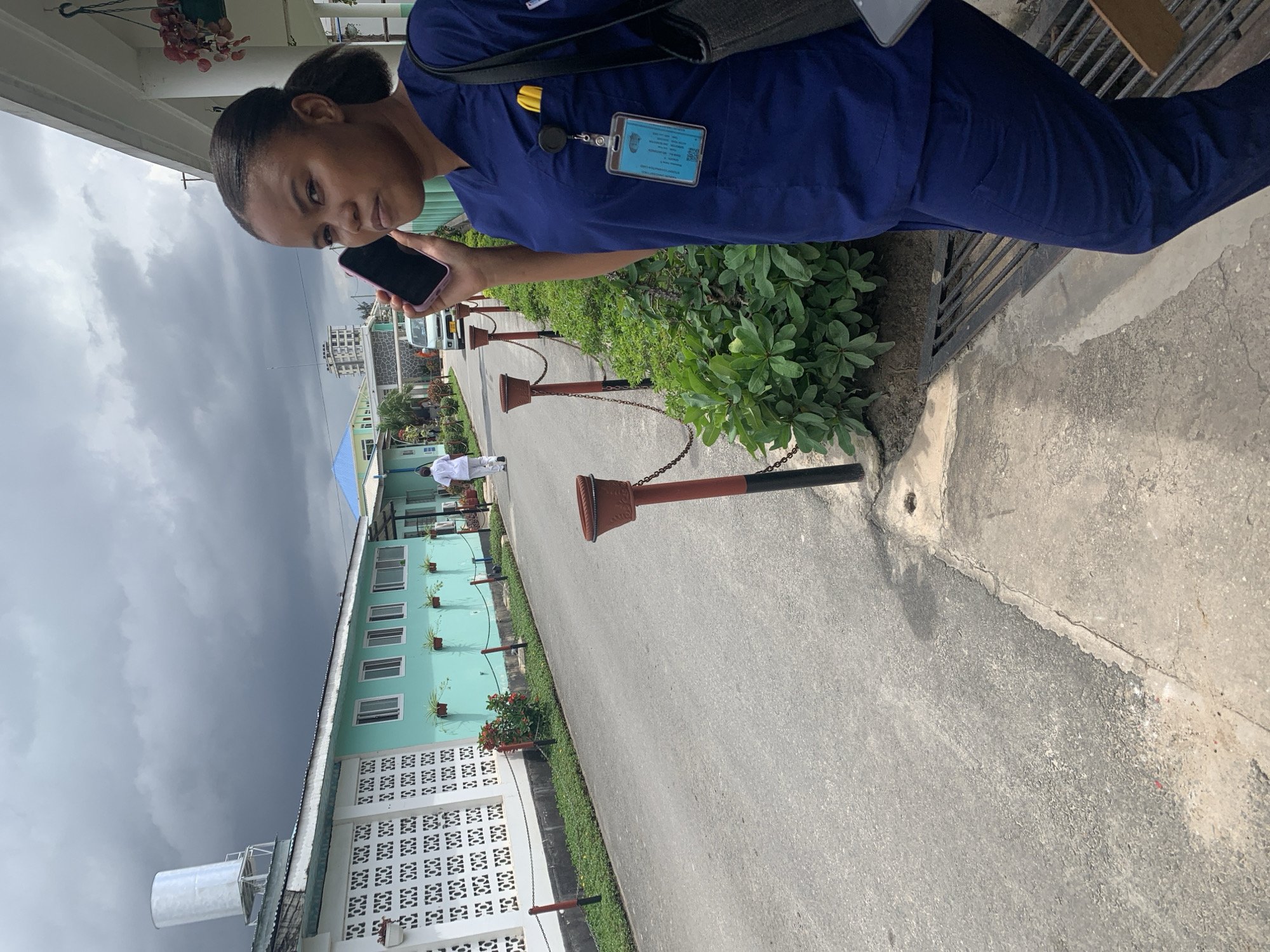 The image captures a woman in navy blue scrubs, seemingly a medical professional, holding a smartphone. She is standing outdoors beside a paved road, adjacent to a building with turquoise and white walls. The building has a distinctive architectural style, with white latticework and potted plants adorning the windows.
A series of brown and black posts connected by chains line the side of the road. Green foliage grows along the edge of the pavement, separating the road from the building. The sky is overcast with dark, ominous clouds.
In the background, a person wearing white clothing is walking along the road. Buildings of varying heights can be seen in the distance. The woman has an ID badge visible on her scrubs.
Based on the architectural style of the buildings, and the general setting, the location might be in Dar es-Salaam, Tanzania.