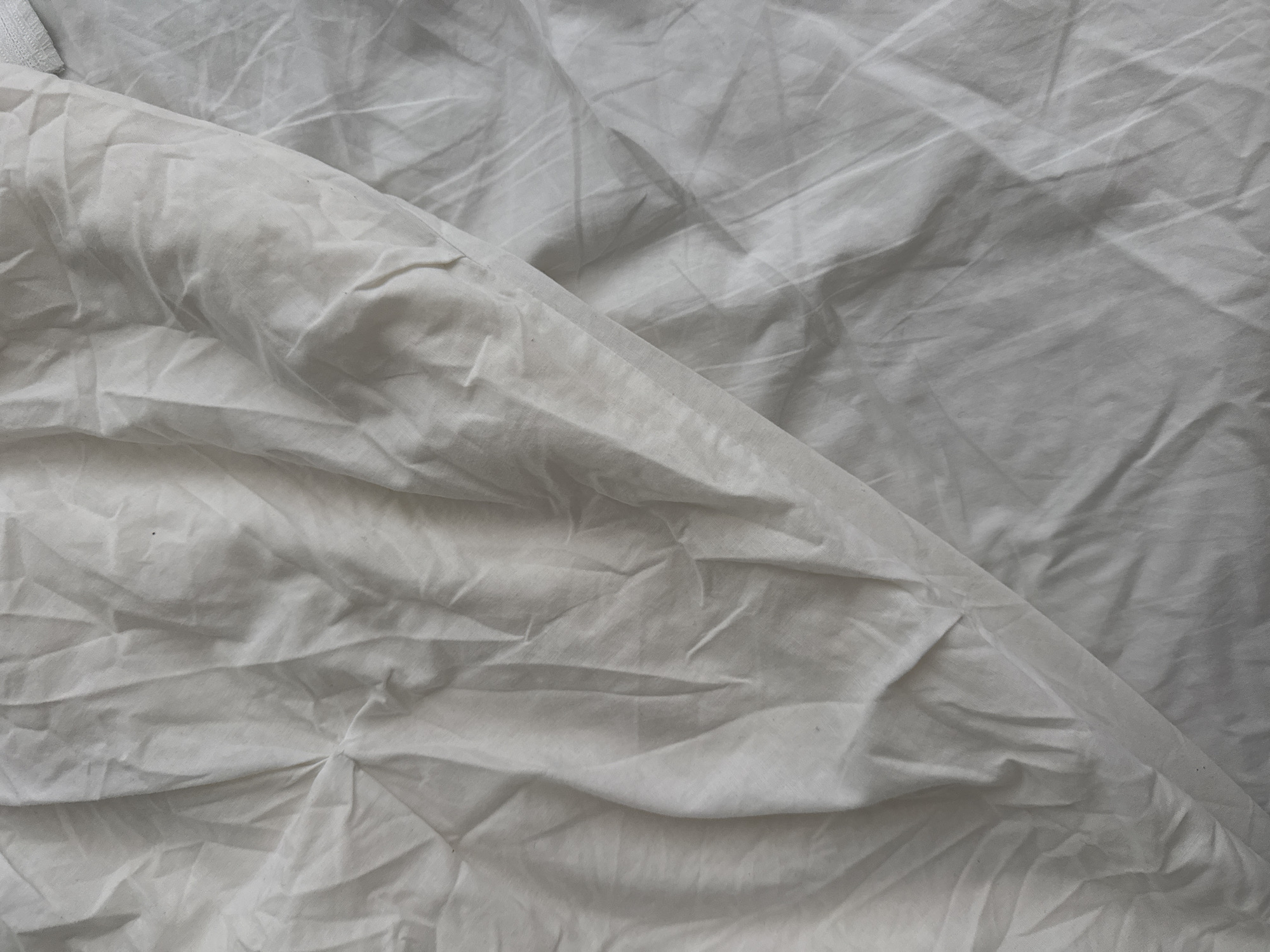 A white fabric lies draped, creating a landscape of shadows and highlights. The material exhibits numerous folds and creases, suggesting it is either crumpled or has been carelessly laid out. The texture of the fabric appears to be relatively smooth, with a slight sheen that catches the light. The overall impression is one of soft, flowing lines and a delicate texture. There are no people, animals, or specific objects visible in the frame. The background is entirely obscured by the fabric itself.