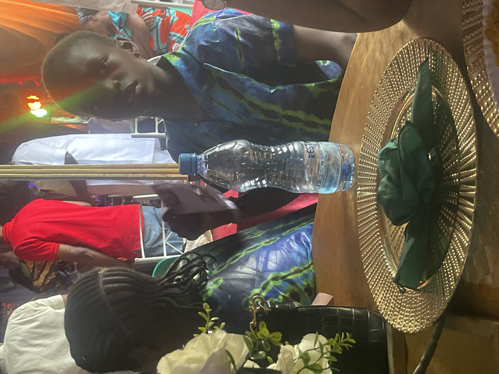 The image shows a gathering of people, possibly at an event, in Jos, Nigeria. A person with a short haircut, wearing a blue and green patterned shirt, is in focus. A bottle of water sits on a table next to a gold-colored plate with a dark green napkin. Another individual in a red shirt is visible in the background. A woman with braided hair is also present near a black purse and white flowers. The setting appears to be indoors with stage lighting visible in the background.
