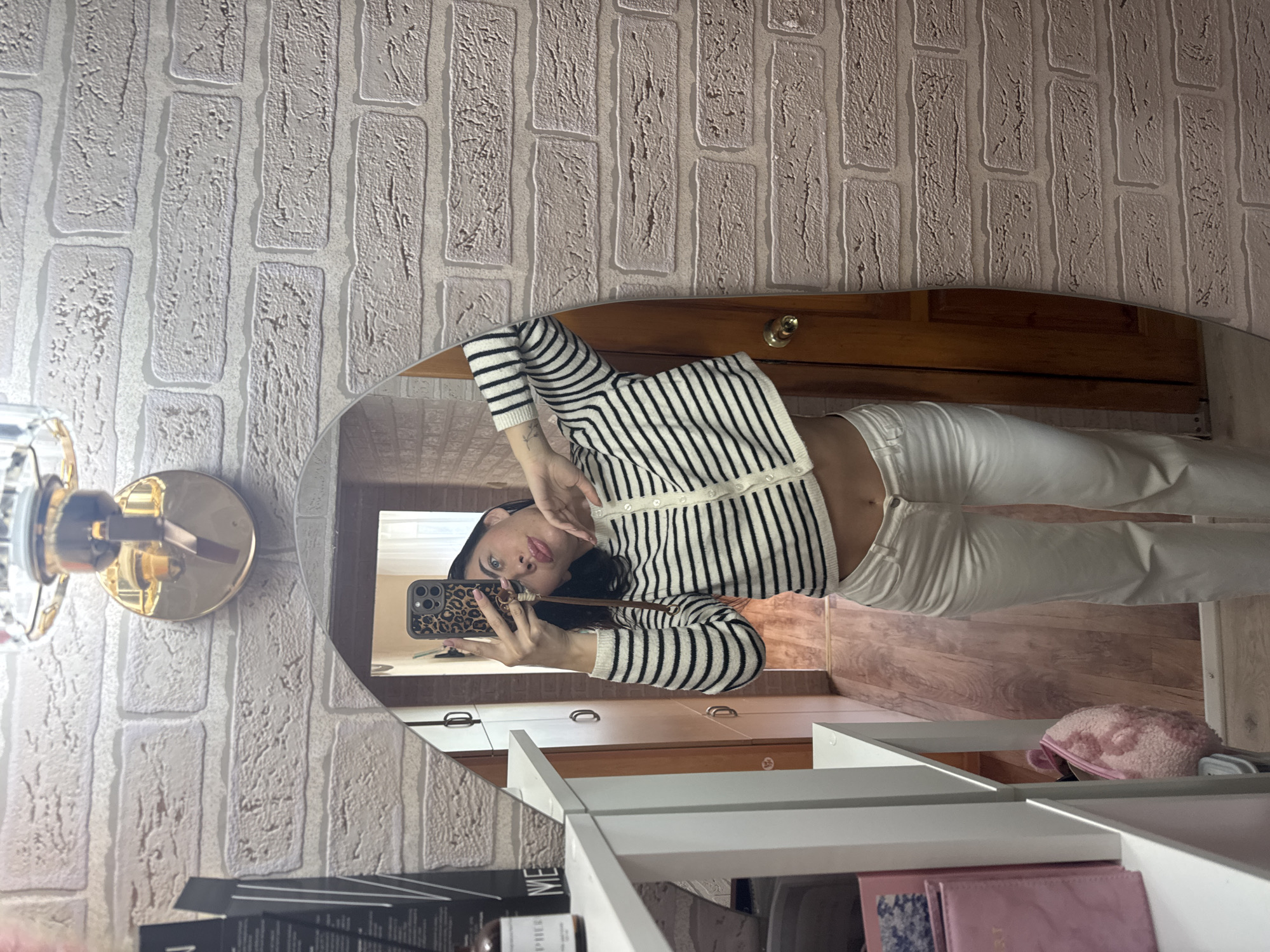 In an indoor environment, a young woman with dark hair, wearing a black and white striped cropped cardigan and white pants, is reflected in a mirror. She is holding a smartphone with a leopard-print case and taking a selfie, puckering her lips towards the camera. Her midriff is exposed. The background behind her reflection shows a wooden door and a hallway. The wall in the foreground has a light-colored brick wallpaper. The overall scene suggests a personal space, possibly a dressing area or bedroom. The image does not contain any information about the location in the city of Chaikovsky, Russia.