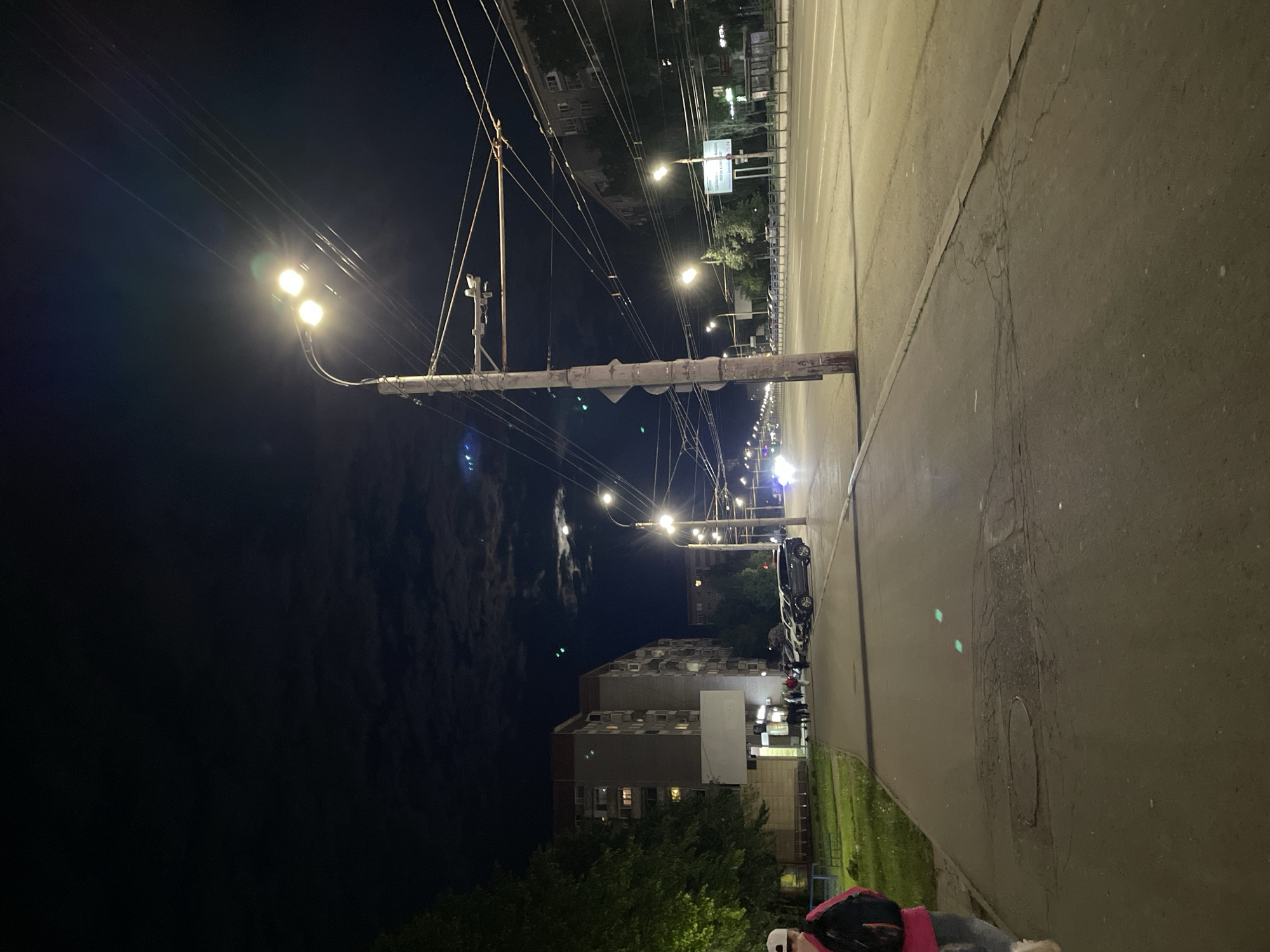 At night in Izhevsk, Russia, a street scene is illuminated by streetlights. A wooden pole supports overhead wires and streetlights. The street pavement is dark and cracked, with a grassy strip and parked cars visible on one side. Buildings are visible in the background. A person wearing a pink backpack and a white helmet is located in the bottom right corner of the image.