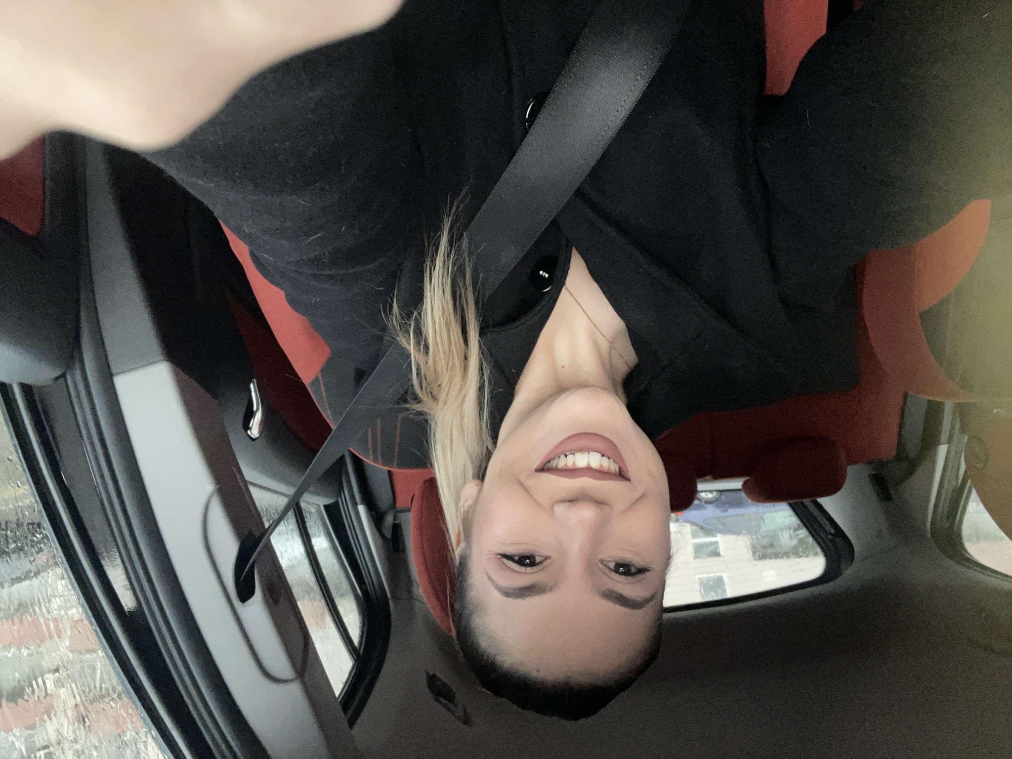 The image features a smiling woman seated inside a car, likely taking a selfie. She has blonde hair pulled back in a ponytail and is wearing a black coat with buttons and a thin, delicate necklace. A black seatbelt is fastened across her chest. Her expression is a wide, closed-mouth smile, showing her teeth and conveying a positive, happy emotion.
The setting is the interior of a vehicle, with parts of red car seats visible as accents against the black upholstery and the car's dark roof. Through the car windows, the outside environment is discernible. Streaks of water on the windows suggest recent rain or condensation. In the background, elements of an urban or suburban landscape are visible, including another blue vehicle and a building with windows.
The activity taking place is a self-portrait within the car. The time of day appears to be daytime, with soft, diffused lighting, possibly due to overcast weather or rain. There is no visible text in the image. The provided context for the location is Burgas, Bulgaria.
