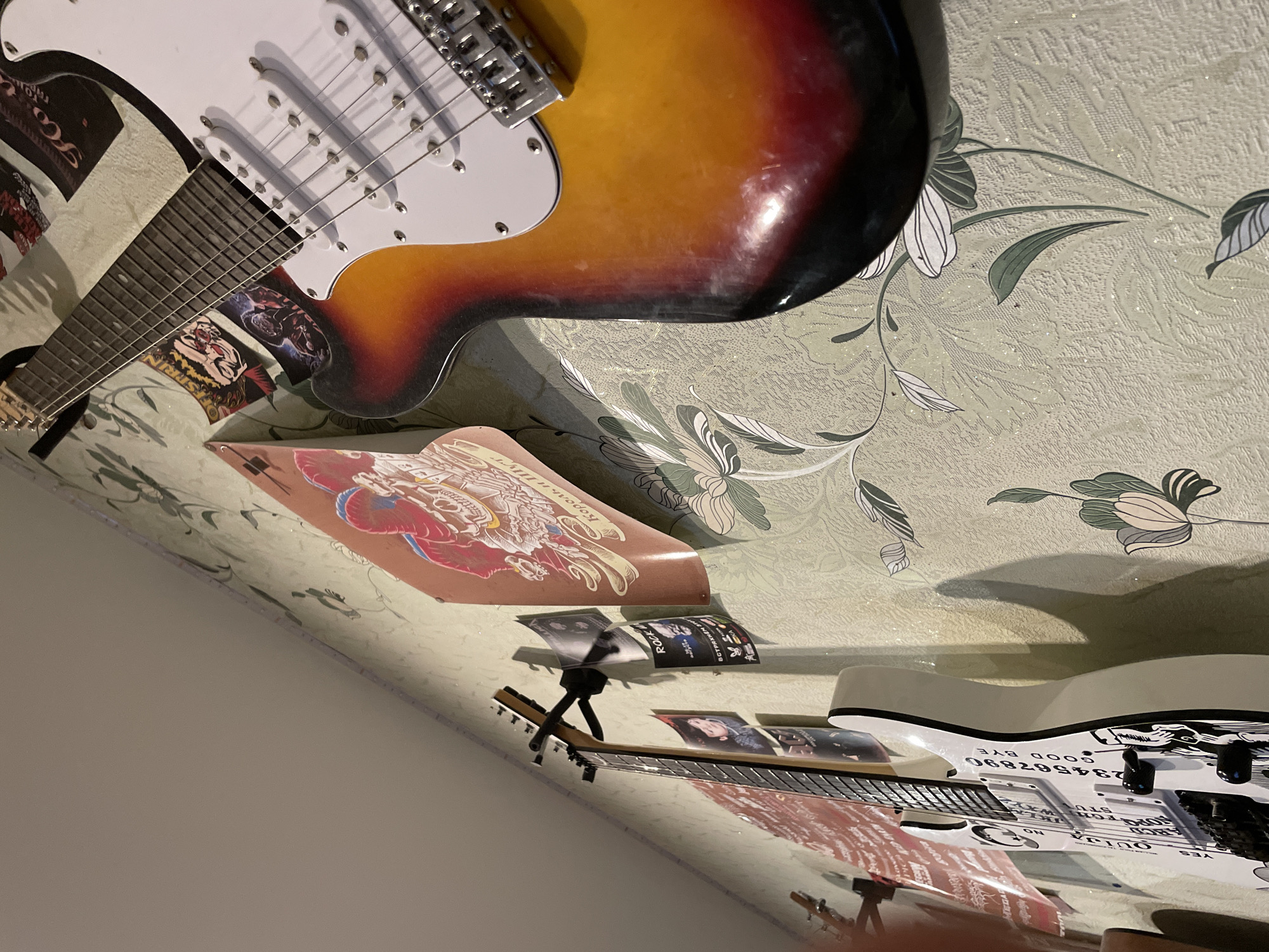 The image, taken in Chaikovsky, Russia, depicts an indoor scene showcasing two electric guitars and various musical memorabilia displayed against a patterned wall. The perspective is tilted, offering a dynamic view of the arrangement.
In the upper left, a sunburst-finished electric guitar, resembling a Stratocaster, is prominent. It features a white pickguard, six strings, and visible bridge hardware. Its fretboard is also clearly seen. Behind and around this guitar are several stickers or prints with graphic designs.
Further down and to the right, a distinctive white electric guitar is displayed, notable for its Ouija board-themed graphics. Text clearly visible on this guitar includes "YES," "OUIJA," "ON," "GOOD BYE," and the numbers "234567890." Parts of its fretboard and headstock are also visible, supported by a stand.
The background wall is covered with light-colored wallpaper featuring an embossed, textured floral pattern of white and dark green leaves and flowers. Numerous prints and posters are affixed to the wall, placed between and around the guitars. One poster prominently displays "Король и Шут" (King and Jester), a well-known Russian punk-rock band, providing a direct link to the location context of Chaikovsky, Russia. Other visible text snippets from these prints include "ROCK," "В СТРЯХНЕM TOT" (possibly "Let's shake that..."), "MEGAR," "SPRIN," "KA," and "Ho."
There is no discernible activity or event taking place; the scene presents a static collection of instruments and fan items. The lighting appears to be artificial indoor light, casting subtle shadows. The image captures a personal space decorated with an evident enthusiasm for music, particularly rock, and Russian punk-rock culture.