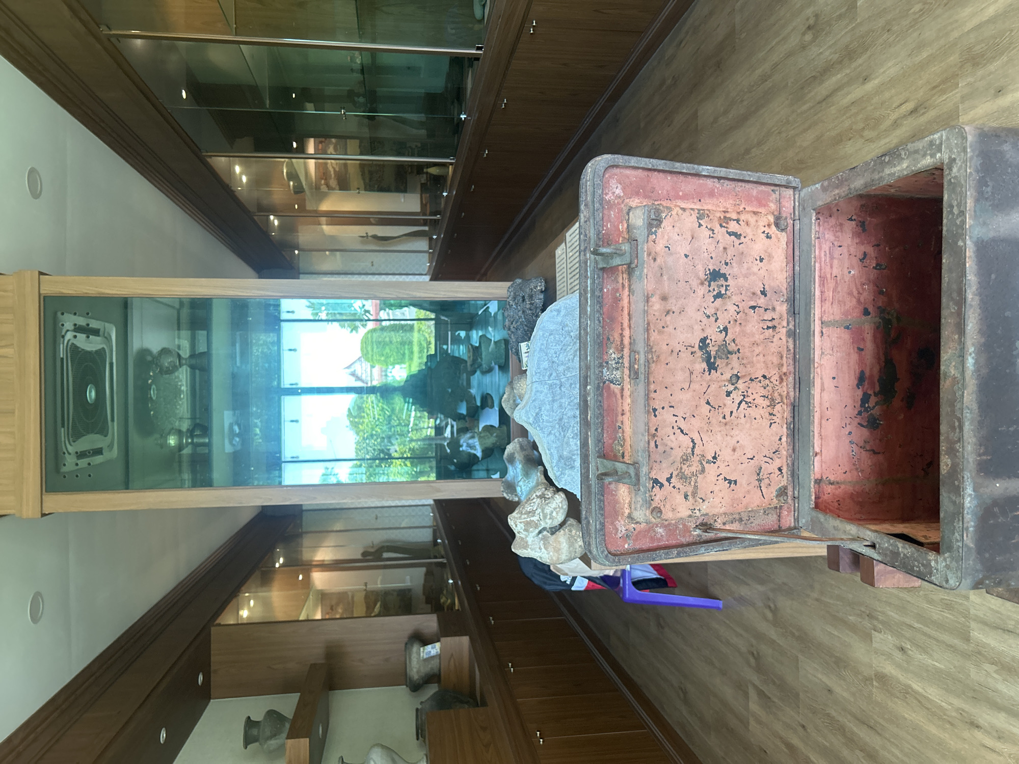 This is a medium shot, eye-level interior photograph of a museum display room. The room features long, wooden display cabinets with glass doors on either side, showcasing various artifacts. In the foreground, an open, weathered metal chest with a reddish-orange interior is prominently displayed. Behind the chest, on a table, are several large, light-colored stones and a dark, textured object. A purple plastic chair with a black bag draped over it is visible to the left of the stones. The room has a light-colored wooden floor and recessed ceiling lights. A large glass pane in the center of the room reflects the exterior environment, revealing trees and a building with a sloped roof. The overall atmosphere is that of a historical or cultural exhibition space. The scene is located in the Thai Ko Subdistrict, Thailand, likely a museum or cultural heritage center.