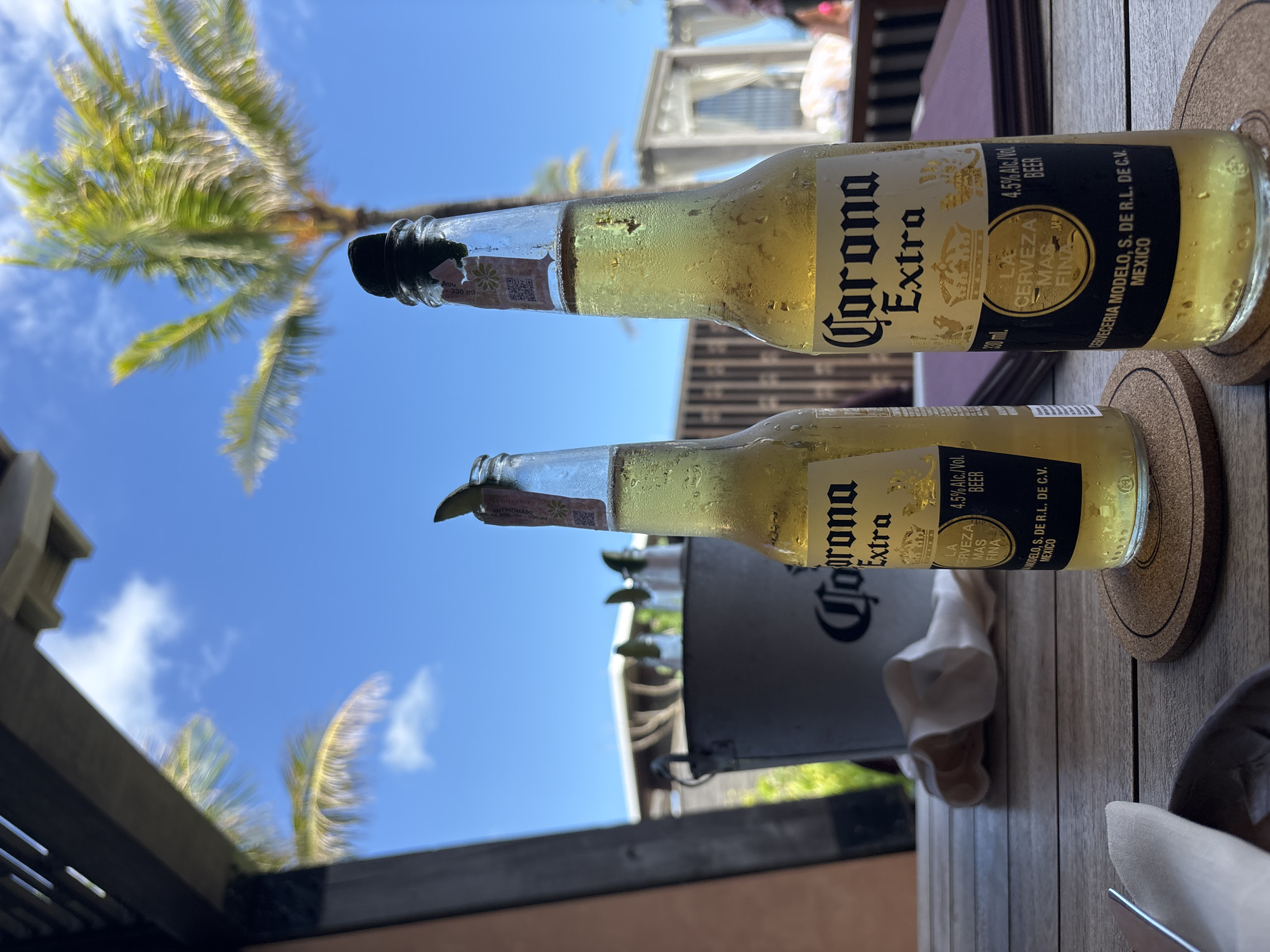 Two bottles of Corona Extra beer sit on a wooden table. The bottles appear partially consumed and have condensation on them. In the background, there is a blue sky, palm trees, and glimpses of buildings, with elements suggesting an outdoor setting. The image possibly captures a scene in a tropical location. The labels on the bottles are clearly visible, indicating the brand, alcohol content (4.5% Alc./Vol), and place of origin (Mexico). There is a coaster under one of the bottles and some other tableware visible.