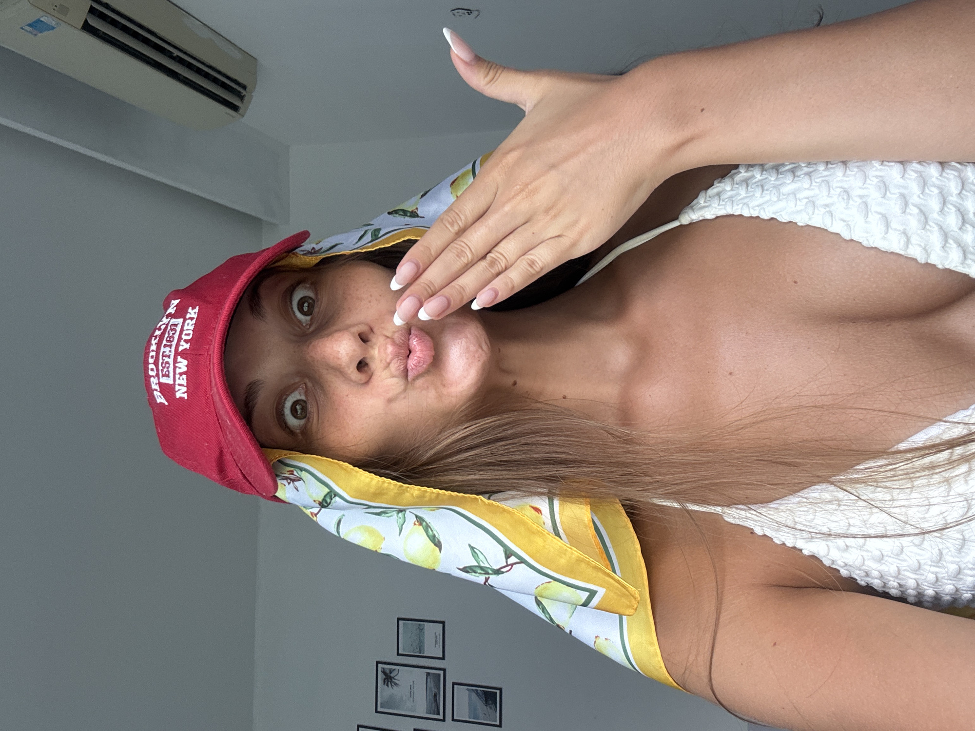 A woman is positioned at an unusual angle, her face central. She is pouting with her lips pursed, her left hand raised in front of her face. She is wearing a red baseball cap with "BROOKLYN EST. 1831 NEW YORK" emblazoned on it. A scarf with a lemon print is draped around her head and neck. She wears a white, textured top. The background includes a white wall adorned with a grid of framed pictures and an air conditioning unit near the ceiling. The location within the city of Da Nang, Vietnam cannot be determined from this image.