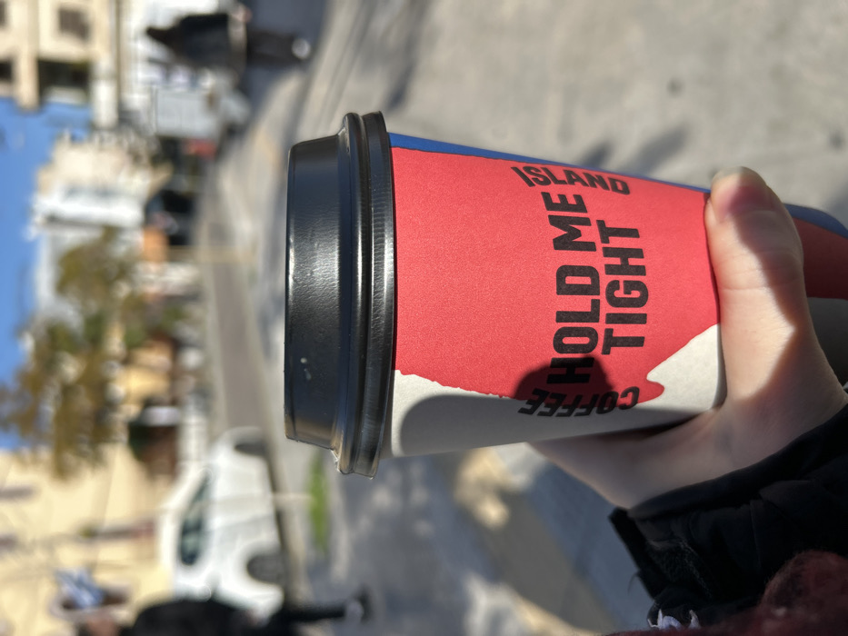 A close-up shot captures a hand holding a coffee cup outdoors on a sunny day in what appears to be Nikea, Greece. The right hand, with fingers wrapped around the cup, is positioned prominently in the foreground. The coffee cup is the central focus, featuring a vibrant design with a red body, a blue band near the rim, and an irregular white shape towards the bottom. A black plastic lid covers the top of the cup.
Bold black text is visible on the cup, reading "ISLAND" and "HOLD ME TIGHT" arranged vertically. Below this, the word "COFFEE" is also visible, printed upside down relative to the other text. Bright sunlight casts distinct shadows across the cup and the hand, indicating clear and sunny weather conditions during the daytime.
The background is heavily blurred, suggesting a shallow depth of field, but hints at an urban outdoor setting. Vague shapes of buildings, some green foliage from trees, and a white vehicle can be discerned, consistent with a street scene. The image portrays a casual moment of someone enjoying a beverage while possibly walking or standing outdoors.
