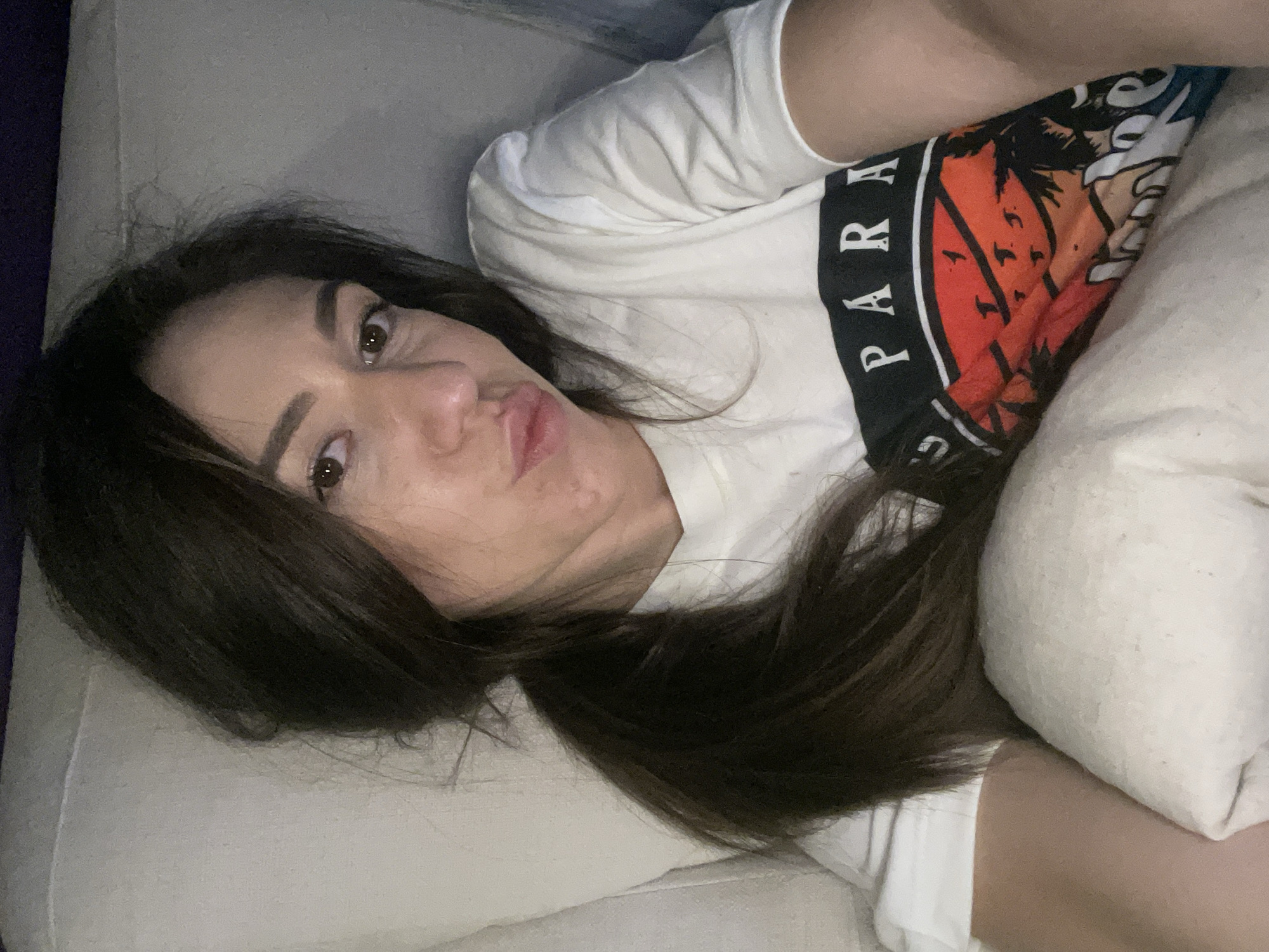 A young woman with dark hair lies on a light-colored sofa or bed. She is wearing a white t-shirt with a graphic print including the word "PARADISE". She is partially obscured by a white pillow. Her face is visible, and she has a slightly puckered expression. The image is taken from an overhead angle. There is no information to identify the location as Sofia, Bulgaria.