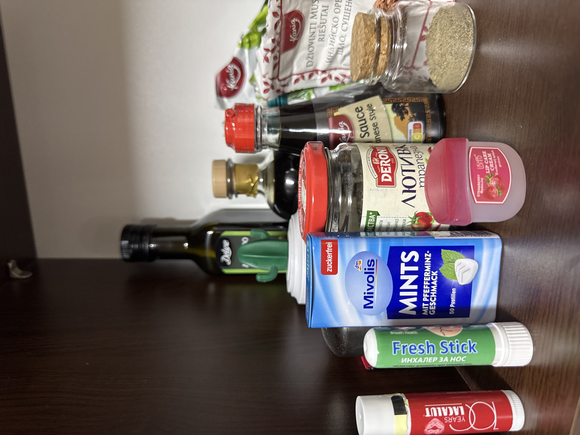 The image shows a cluttered shelf containing various food items and personal care products. Visible are items such as spices in glass jars, a bottle of olive oil labeled "Deluxe", a bottle of Japanese-style sauce, a jar of Deroni lutenica, Mivolis mints, a Fresh Stick nasal inhaler, and Lacalut lip balm. There is also a bag of dried spices labeled with Bulgarian text, and a small lip care cream. All items are arranged haphazardly on what appears to be a dark brown wooden shelf. The scene is most likely set in Bulgaria, specifically in Sofia, based on the Bulgarian text present on several of the products.