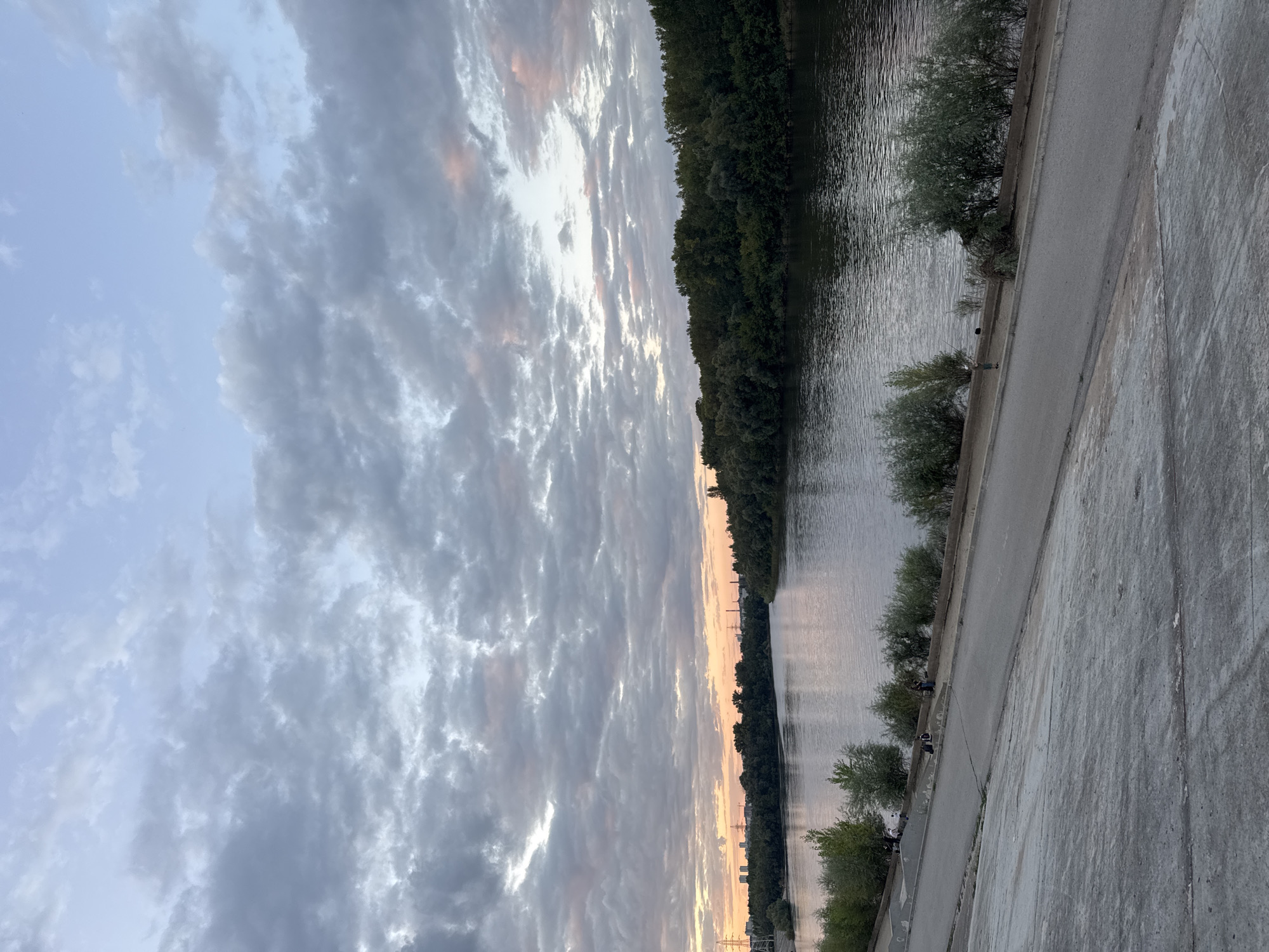The image captures a serene riverside scene in Ufa, Russia, likely during twilight. The foreground is dominated by a sloping concrete embankment, a common feature along the embankment of the Belaya River, a significant waterway in Ufa. The concrete is textured and shows signs of wear, with visible cracks and lines indicating sections.
Beyond the embankment, a paved pathway runs parallel to the river. Several individuals are present along this pathway and on the grassy area between the path and the water's edge. Some are seated, appearing to relax or fish, while others are standing. One person is visibly holding a fishing rod, suggesting recreational fishing. Scattered bushes and small trees dot the landscape along the riverbank, adding to the natural setting.
The Belaya River itself occupies the midground, its surface reflecting the soft, diffused light of the sky. The water appears calm, with gentle ripples. Across the river, a dense line of trees forms a dark green backdrop, characteristic of the riverside vegetation found in and around Ufa. In the far distance, silhouetted against the horizon, are the structures of the city, including tall buildings and industrial elements like power lines and towers.
The sky above is filled with a dramatic display of clouds. A mix of gray and white clouds stretches across the frame, with patches of soft pink and orange light peeking through in the upper right quadrant, indicating the lingering glow of sunset or the approaching dawn. The overall atmosphere is tranquil and contemplative, evoking a sense of peace at the end of the day.