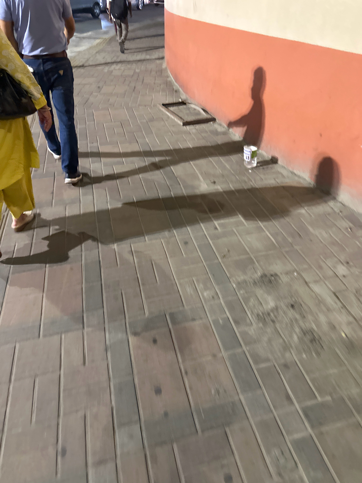 The image shows a paved walkway with a textured surface, likely made of bricks or tiles, in an urban outdoor setting. The walkway is illuminated by artificial light, casting distinct shadows on the ground and a nearby coral-colored wall. In the foreground, the lower legs and feet of two individuals are visible. One person wears a yellow garment and beige footwear, while another wears dark blue jeans and dark shoes. A dark bag is partially visible on the left. In the background, a person wearing a light blue shirt, dark belt, and blue jeans is walking away from the camera, accompanied by a person in dark clothing with a backpack. The shadow of a third individual, appearing to be a child, is cast on the wall, holding what looks like a plastic bottle. The environment suggests a public space in Kathmandu Metropolitan City, Nepal, possibly a pedestrian area or a sidewalk adjacent to a building. The lighting and shadows indicate it is likely evening or night.