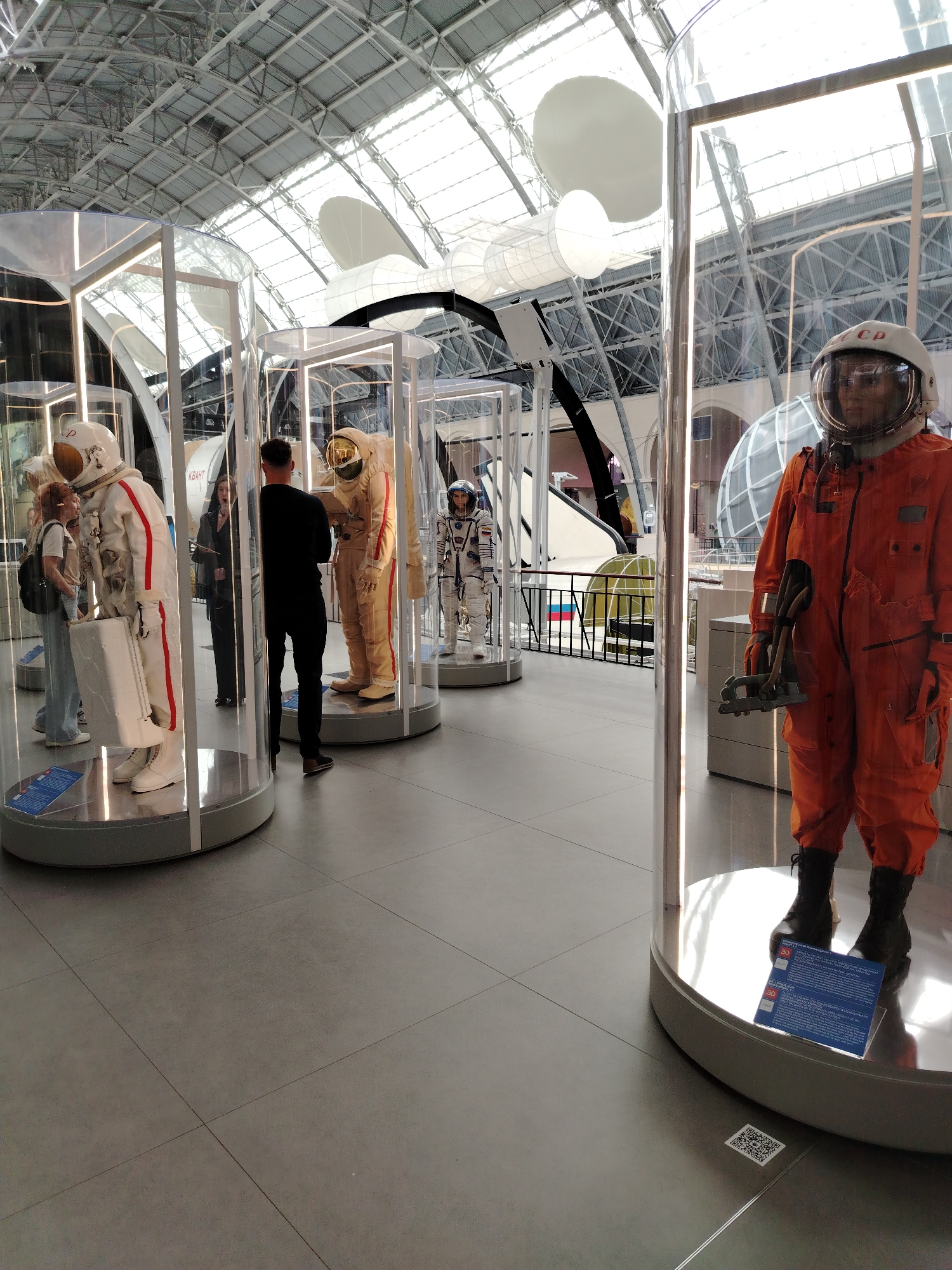In a spacious, modern museum hall with a high, arched, glass-paneled ceiling, several full-body astronaut suits are displayed in cylindrical glass enclosures. The suits vary in color and design, with some being white with red stripes and others being a tan or orange hue. One prominently displayed orange suit in the foreground features a helmet with a clear visor, revealing a mannequin's face. The mannequin is holding a metallic tool. The floor is tiled in a gray, grid pattern. Several visitors are present, observing the exhibits. One woman with red hair is looking at a white suit, accompanied by another person. A man in a black shirt and pants stands facing a tan suit. In the background, more exhibits and architectural features of the museum are visible. The overall impression is that of a museum dedicated to space exploration. This scene is located at the Cosmonautics Memorial Museum in Moscow, Russia.