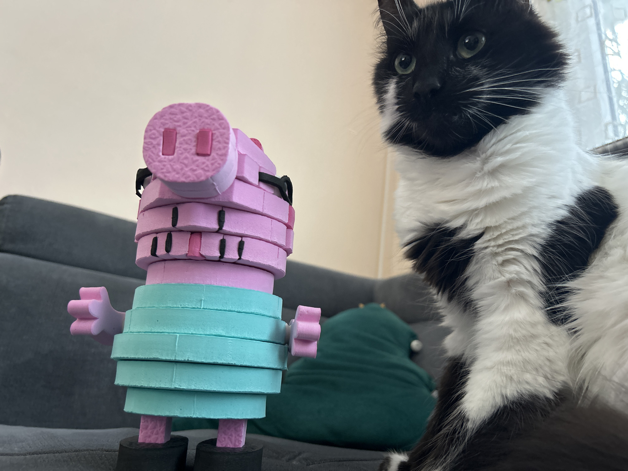 A black and white cat is in the foreground, looking to the left. To the left of the cat is a Peppa Pig-like toy figure made of stacked foam rings. The figure is pink with a blue torso, and stands on a gray couch. A green pillow sits behind the Peppa Pig figure. The background is out of focus, but includes a wall and window. The image does not contain enough information to determine the location within the city of Plovdiv, Bulgaria.