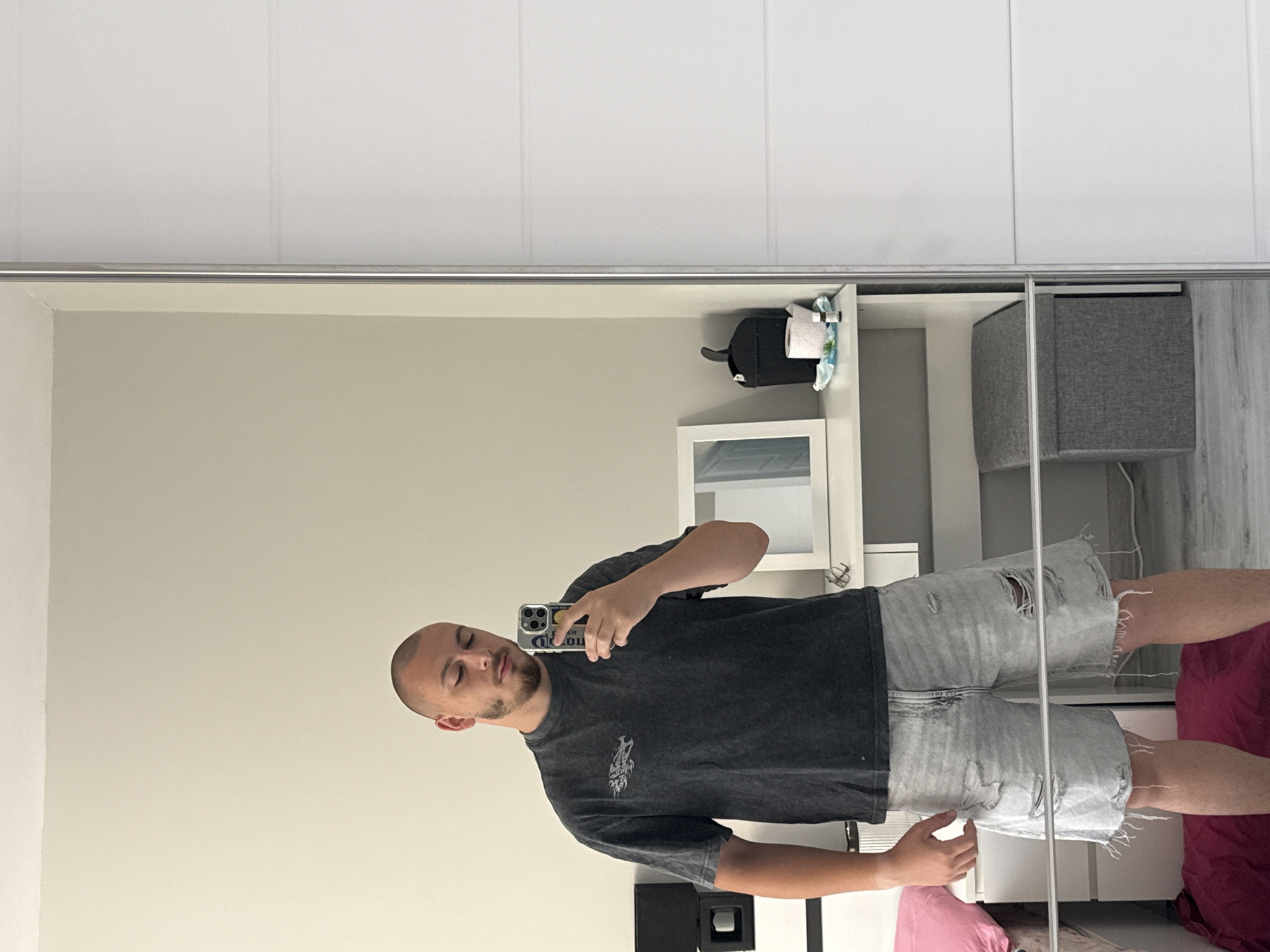 A young man with a shaved head and a beard is standing in front of a mirror, taking a selfie with his phone. He is wearing a dark gray, distressed t-shirt with a graphic on the left chest and light-wash, ripped denim shorts. He is positioned in a bedroom, with a dresser and a large mirrored wardrobe visible. A pink pillow is on a bed to the left, and a dark red blanket is on the floor. A white shelf unit is behind the man, holding a framed picture, a black trash can, toilet paper, and a small bottle. A gray storage ottoman is on the floor in front of the wardrobe. The environment suggests a domestic setting within an apartment in Stara Zagora, Bulgaria.