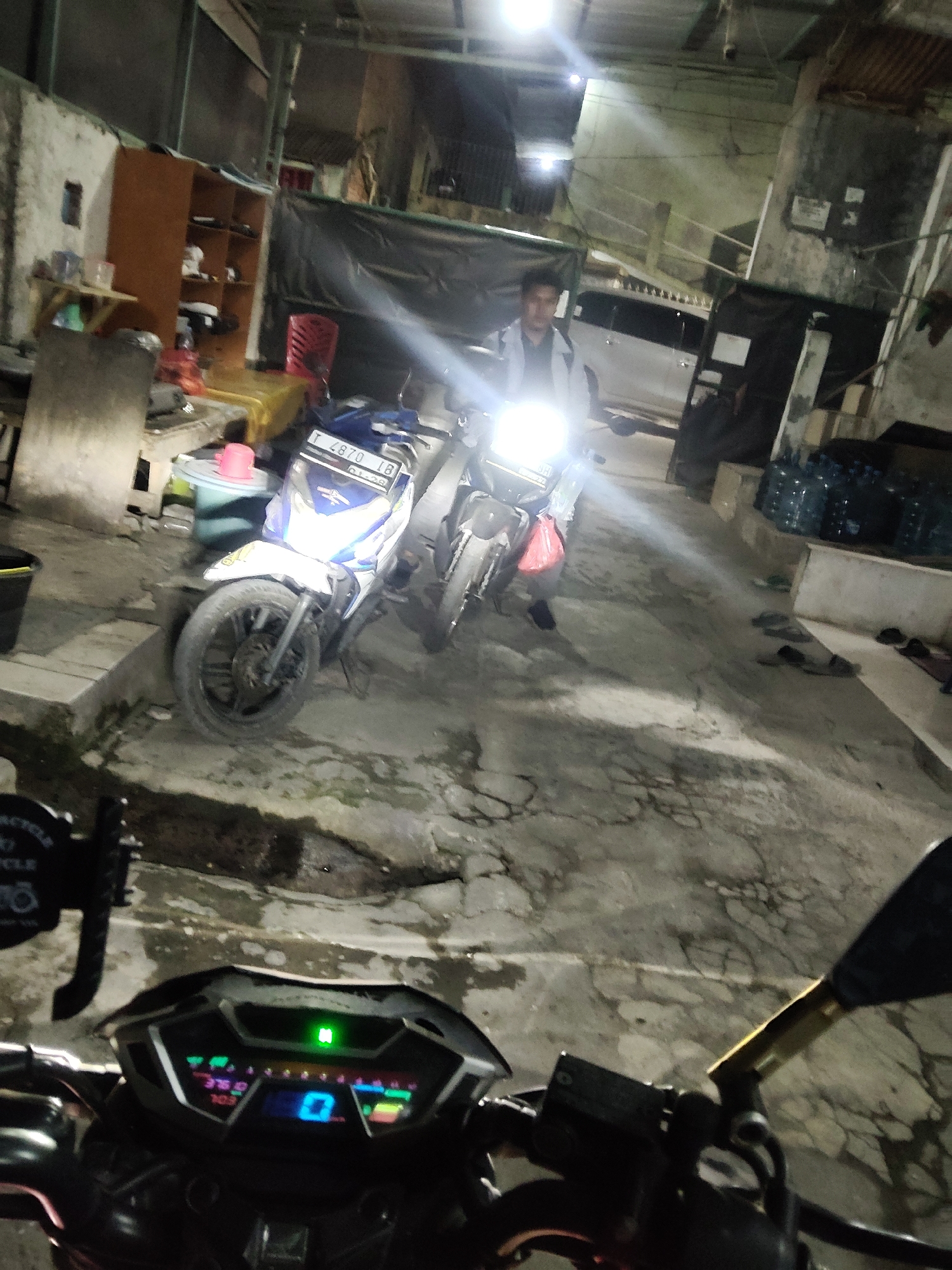 The image depicts a nighttime scene in what appears to be a storage area or a back alley in Mekarmukti, Indonesia. In the foreground, the dashboard of a motorcycle is visible, displaying a digital readout with illuminated green and blue segments, indicating it is powered on. The handlebars and mirrors of this motorcycle are also present. Further into the scene, two motorcycles are parked. The motorcycle in the center has its headlight illuminated, casting a bright light. A person wearing a dark shirt and a light-colored jacket is seated on this motorcycle. The background features a building with exposed beams and corrugated metal roofing, and several large blue water bottles are stacked on a raised platform. The ground is a concrete surface with visible cracks and puddles. The overall atmosphere is dimly lit, suggesting it is night or a poorly lit indoor space.