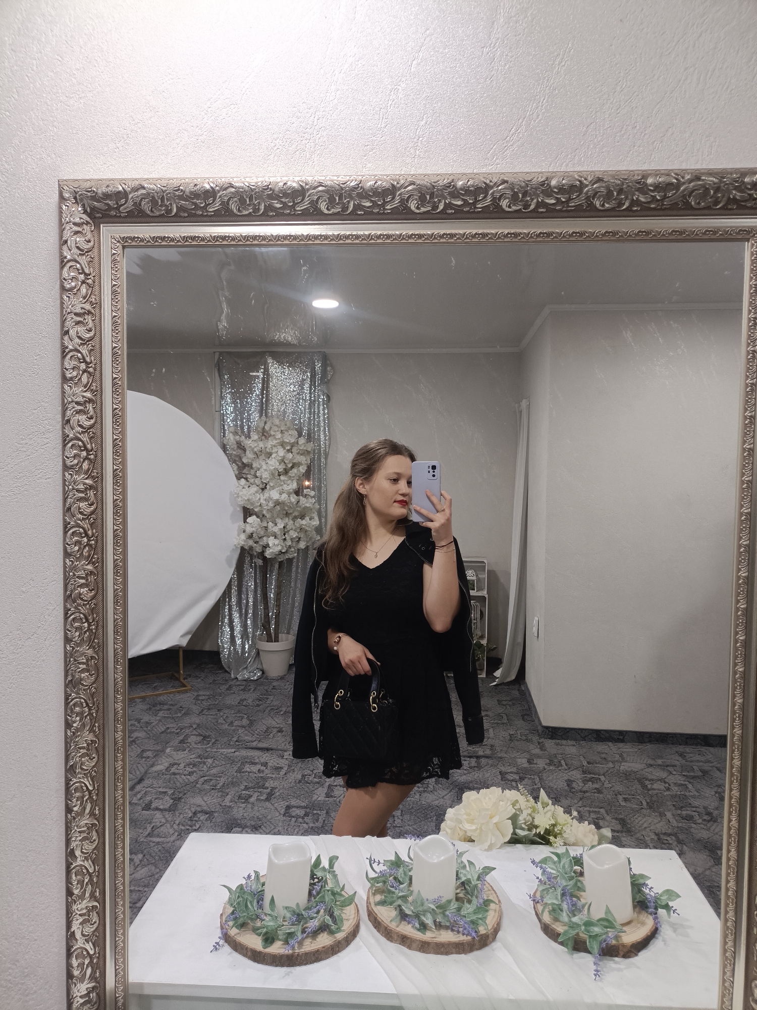 A young woman is taking a selfie in a mirror. She is wearing a short black dress, a black jacket draped over her shoulder, and holding a black handbag. The mirror has an ornate silver frame. The background includes a white backdrop, a sparkly silver curtain, and a white tree. In the foreground, in front of the mirror's reflection, are three white candles on wood slices surrounded by greenery and lavender sprigs. There are also white flowers. The location is likely a studio or event space, possibly in Plovdiv, Bulgaria given the implied prompt context. The woman is looking at her phone and appearing to take a photo of herself.
