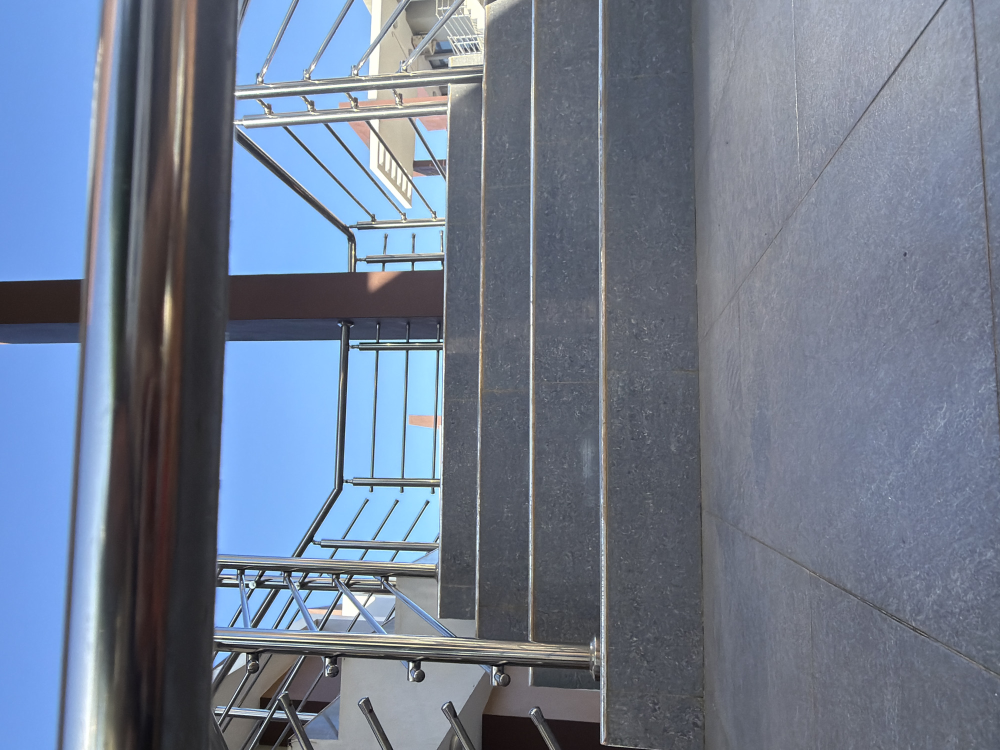 The image shows a view from a low angle looking up a staircase. The stairs are made of gray stone. A stainless steel railing runs along the left side of the stairs. The sky is visible between the railing and the stairs. To the right of the stairs is a gray tiled wall. The location is Unknown, Unknown.