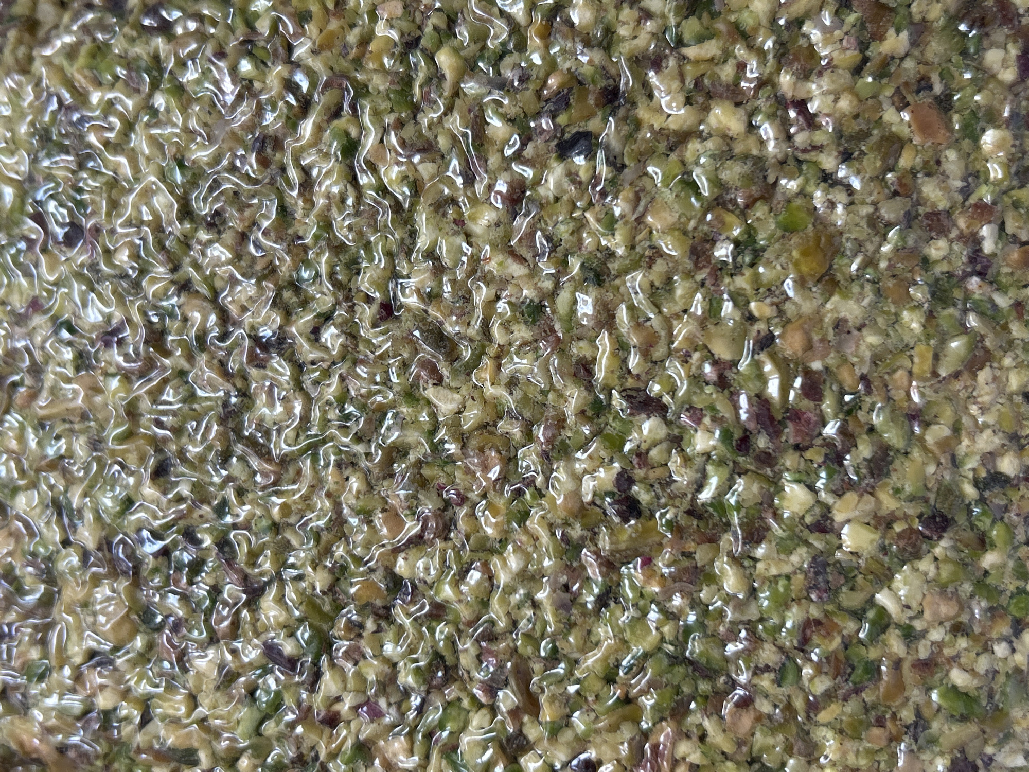 The image shows a close-up of a surface covered in small, granular pieces, predominantly green and brown in color. The surface appears to be coated with a shiny, translucent substance, possibly a glaze or liquid. The individual pieces vary in size and shape, creating a textured and uneven surface. The composition suggests a food-related context or a material made up of crushed components. There is no indication of location.