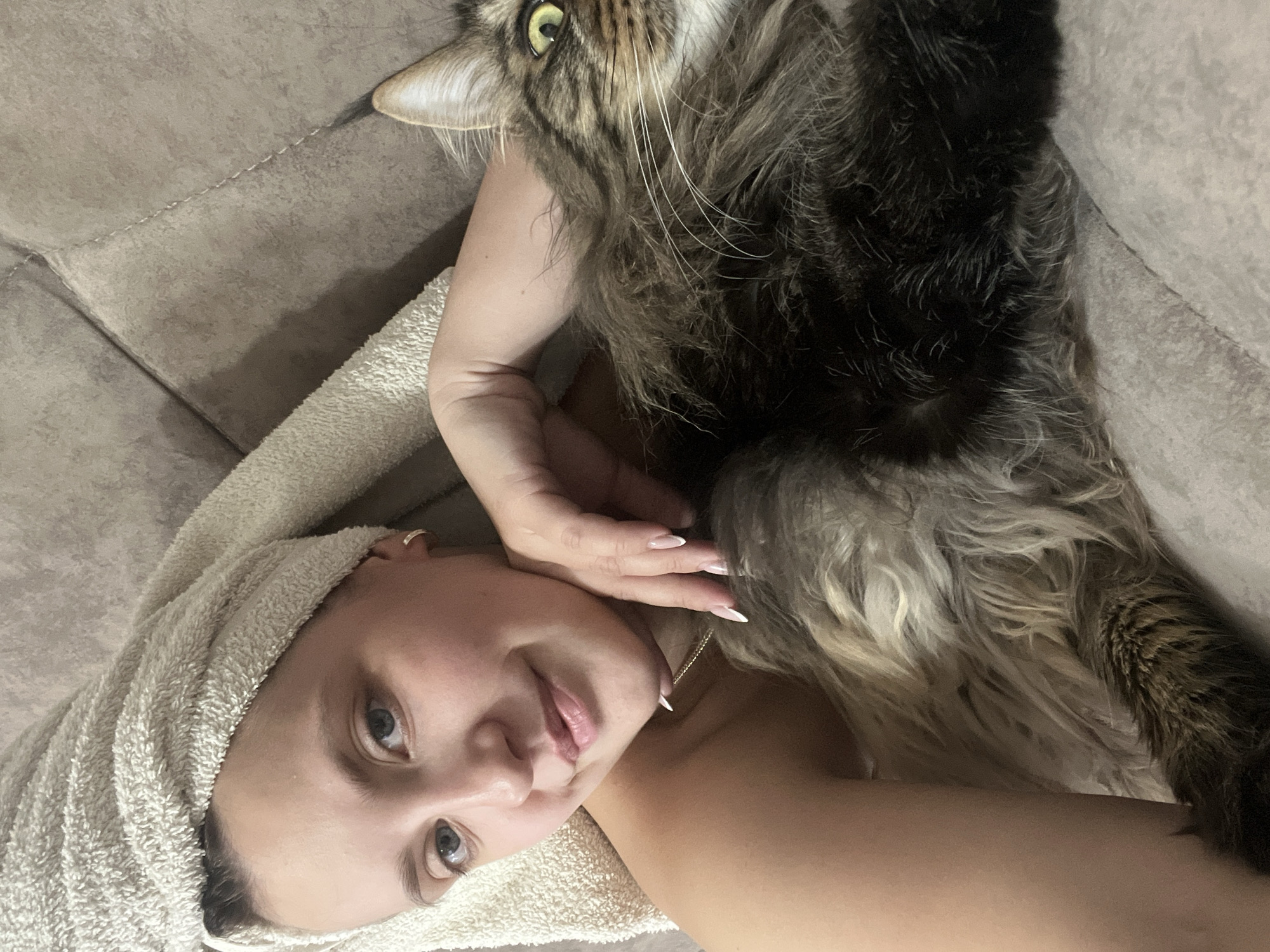 A close-up shot shows a person lying down with a cat on top of them. The person is wearing a towel wrapped around their head and has their eyes open, looking at the camera. Their lips are slightly parted, and they have a gentle smile. A gold necklace is visible around their neck. The cat, a Maine Coon, is lying on the person's chest and abdomen, with its head near the person's face. Its fur is long and striped, and its eyes are wide and green. The person's hand is resting on the cat's fur. The background appears to be a soft, textured surface, possibly a couch or bed. The lighting suggests it is indoors, possibly in the evening or a dimly lit room. There is no visible text in the image. The overall mood is intimate and relaxed, highlighting the bond between the person and their pet.