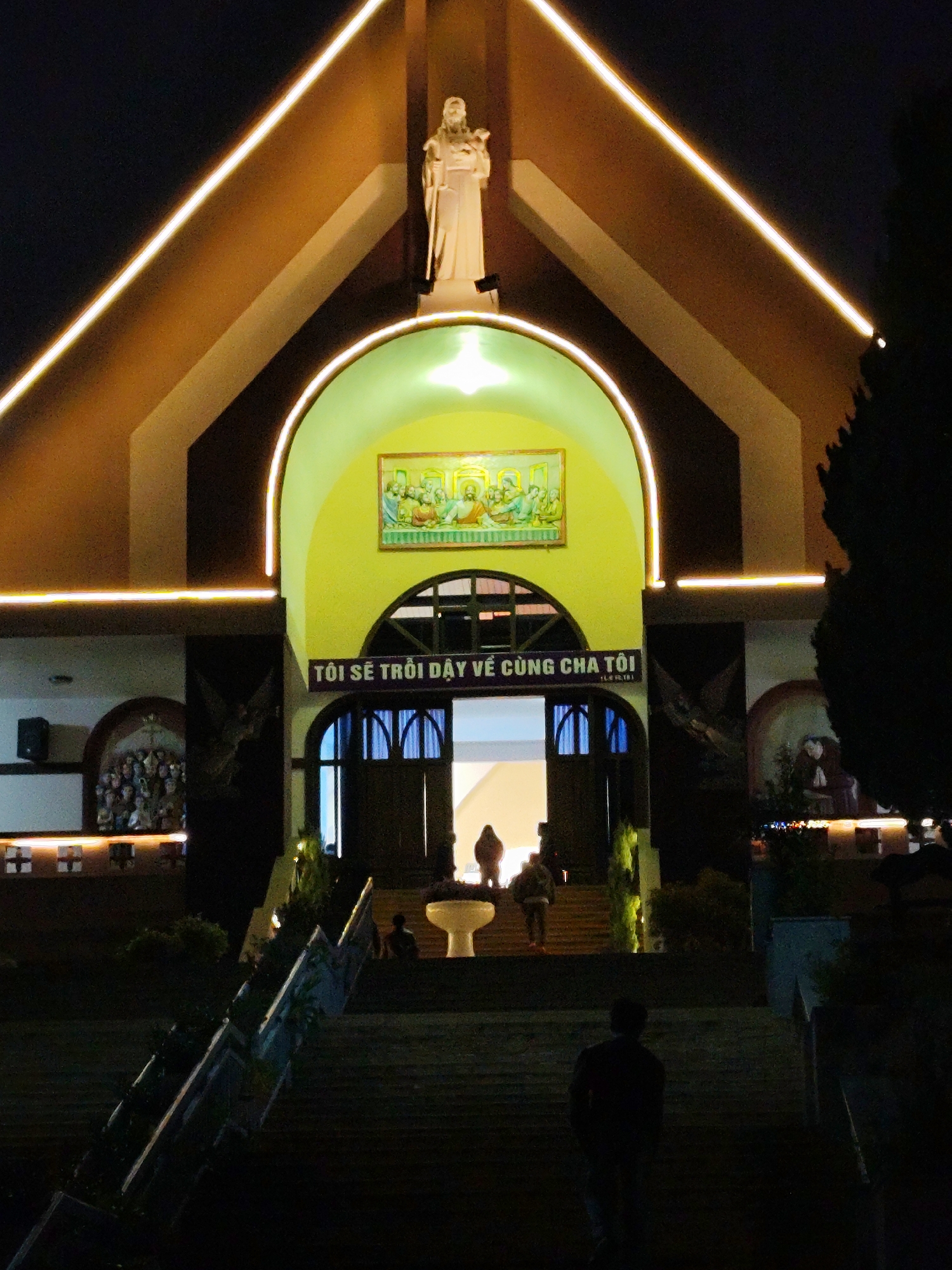 The image displays the illuminated facade of Dalat Cathedral, located in Dalat, Vietnam, during nighttime hours. The central feature is a large, pointed arched entrance with a bright yellow interior soffit. Above this arch, a prominent, light-colored statue of a robed male figure, identified as Jesus the Good Shepherd, stands holding a staff and a lamb. Linear light strips outline the building's gables and the main arch. Within the yellow soffit, a rectangular artwork depicts multiple figures, possibly a biblical scene. A banner with the Vietnamese text "TÔI SẼ TRÔI DẬY VỀ CÙNG CHA TÔI" (I will rise with my Father) is positioned directly above the open, dark wooden entrance doors, which reveal a brightly lit interior. To the left of the main arch, an arched alcove contains a sculptural group of figures, with a black speaker mounted above. To the right, another arched alcove features a single kneeling figure. A wide, multi-tiered stone staircase, lined with decorative railings and sparse vegetation, leads up to the cathedral entrance. In the foreground, one individual, seen from behind, ascends these stairs. Several other individuals are visible in silhouette standing within the brightly lit doorway and on the steps leading into the building, indicating entry or exit activity. The scene is predominantly dark, with the cathedral's artificial lighting providing the primary illumination.
