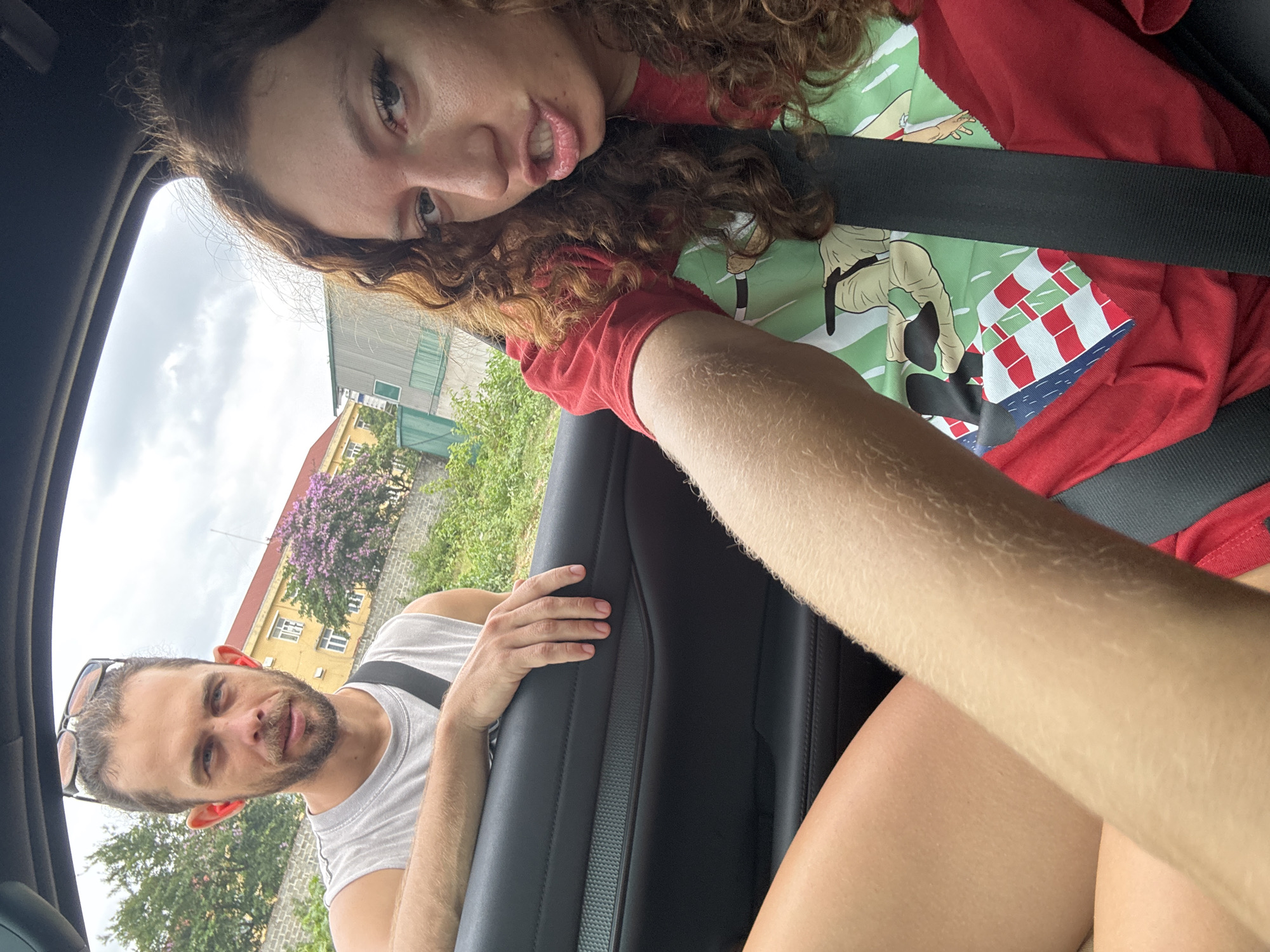 A selfie taken inside a vehicle, presumably a car, shows two individuals. The person in the foreground, a woman with curly brown hair, fair skin, and wearing a red t-shirt with a cartoon print, is angled towards the camera. She is wearing a seatbelt. Behind her to the left is a man with dark hair, a beard, fair skin, and sunglasses perched on his head. He is wearing a light-colored tank top and also has a seatbelt on. His hand rests on the car door. The car is parked outside. Outside the car's window, there is a building, a lilac tree, and other green vegetation. Based on the surrounding environment, the location appears to be in Trung Tam Ward, Vietnam.