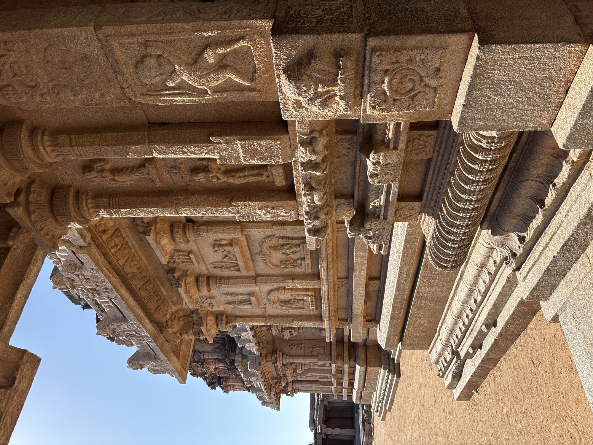 The image shows the underside of a stone structure, likely a temple, in Kamalapura, India. The structure consists of multiple layers of intricately carved stone beams and pillars. The carvings depict various figures, floral patterns, and geometric designs. The stone is a light brown color. The sky is visible in the lower left corner, indicating that the photograph was taken during the day. The overall perspective suggests the photo was taken from a low angle, looking up at the structure.
