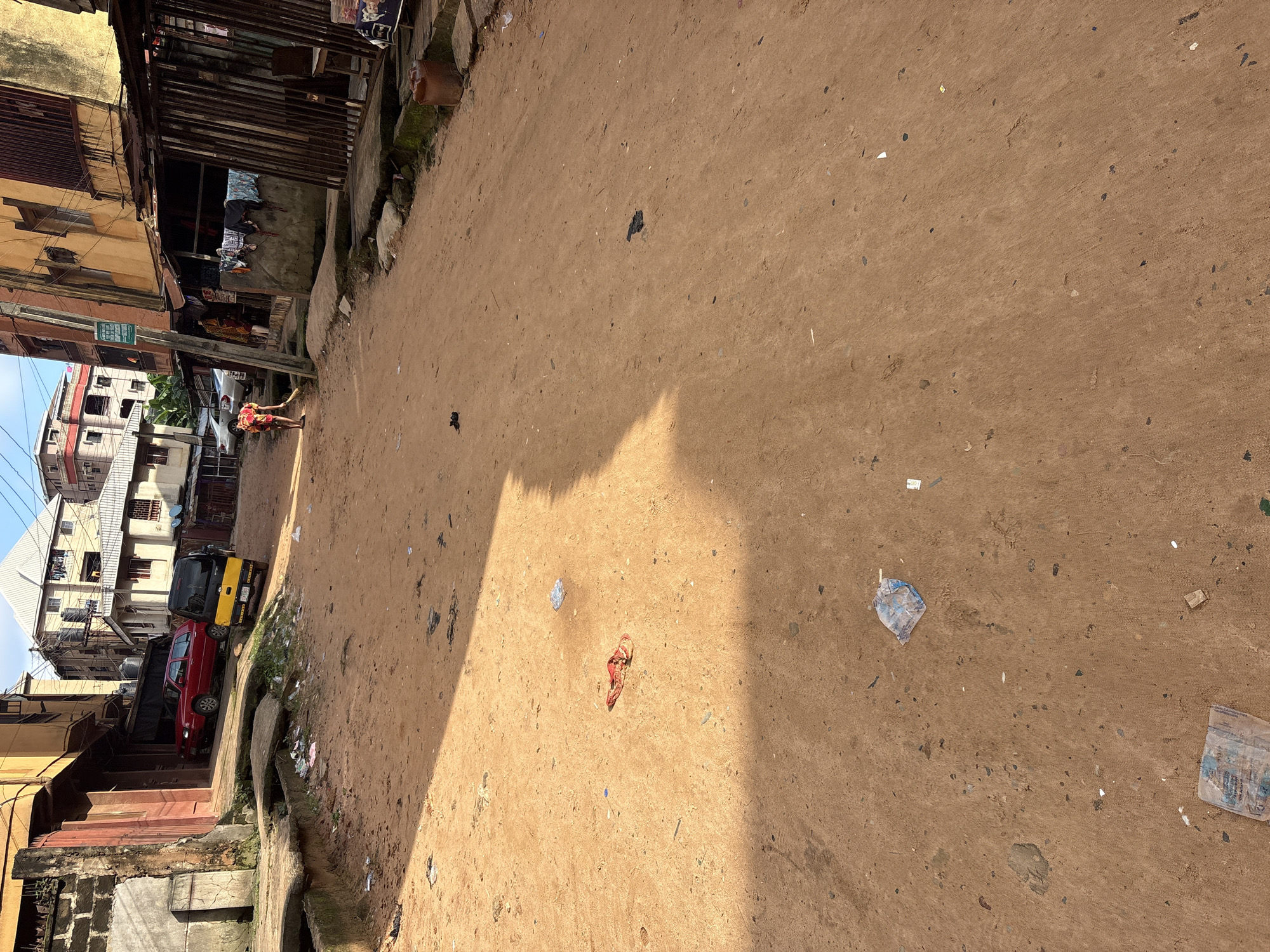 The image depicts a dirt road in Onitsha, Nigeria, lined with multi-story buildings and open-fronted shops. The road surface is composed of reddish-brown earth, uneven and littered with debris, including plastic bags and other assorted trash. In the foreground, a small pile of red fabric lies on the ground, illuminated by a strong shadow from the sun. Further down the road, a person is seen sweeping the dirt with a broom. Vehicles, including a red car and a yellow and black tricycle, are parked on the side of the road. The buildings exhibit a mix of architectural styles and states of repair, with some featuring corrugated iron roofs and others concrete structures. Clothes are hung to dry outside some of the shops, suggesting residential and commercial activity. The overall impression is one of a busy, albeit somewhat unkempt, urban environment.